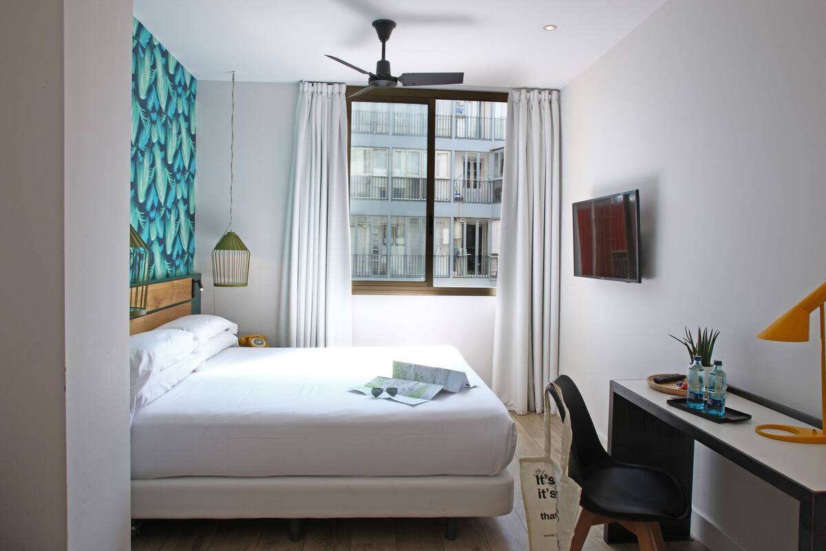 A bedroom with a bed, a desk, a chair, a ceiling fan, and a window with curtains.