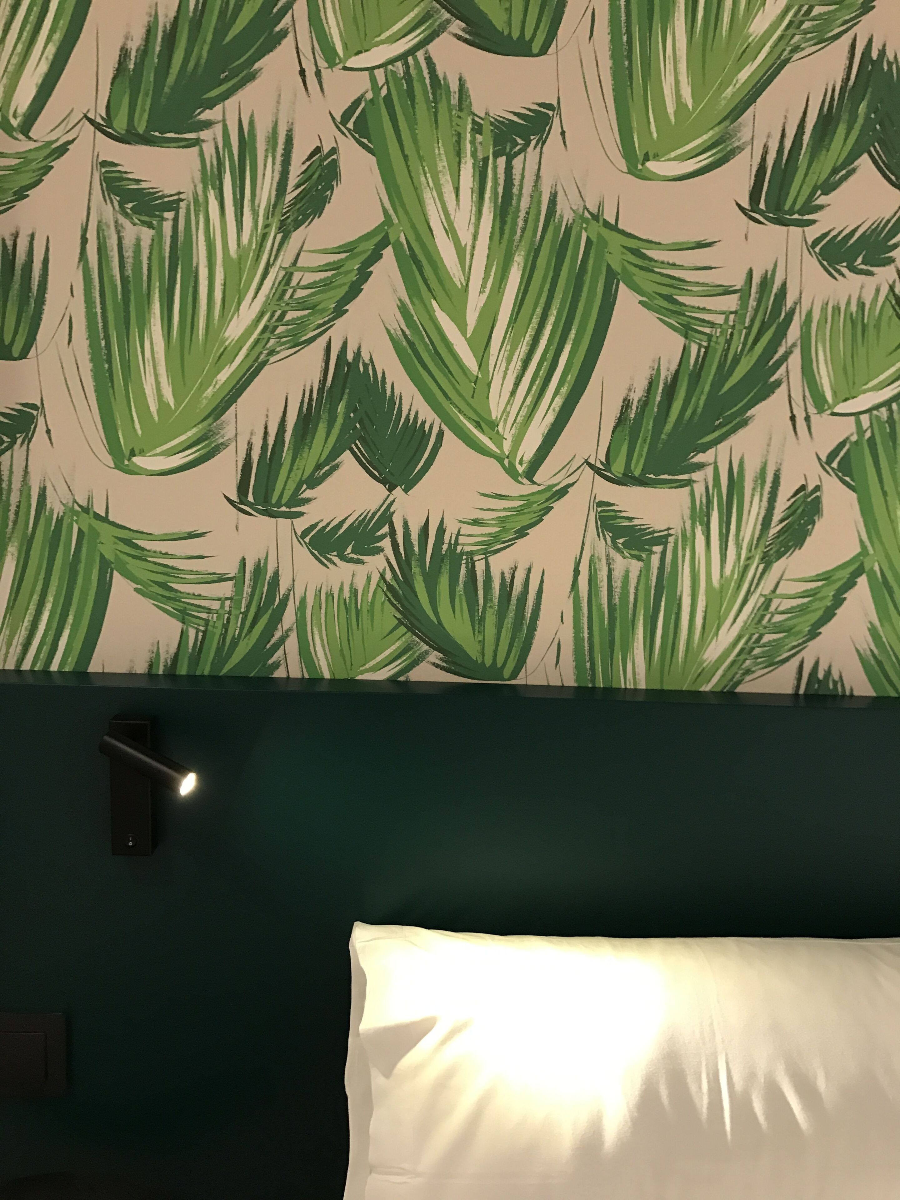 Una habitación con pared verde, papel tapiz estampado, una lámpara de noche y almohada blanca.