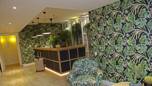 A reception area with a wooden floor, a patterned armchair, a desk with a potted plant, and a wall with a leafy wallpaper design. - chic&basic Lemon Boutique Hotel (Barcelona)