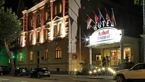 Un hotel con una fachada clásica, un letrero que indica recepción las 24 horas y varias banderas.