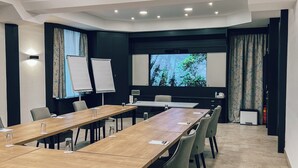 Un salón de conferencias con una larga mesa, sillas, una pizarra, una pantalla de proyector y un amplio ventanal con cortinas.