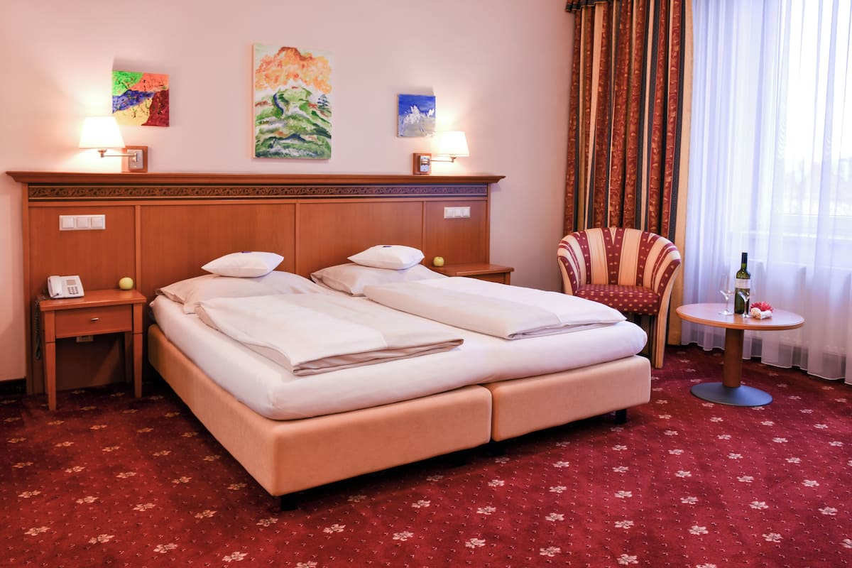 A hotel room with a double bed, two bedside tables, a chair, a small round table with a bottle, and artwork on the walls.
