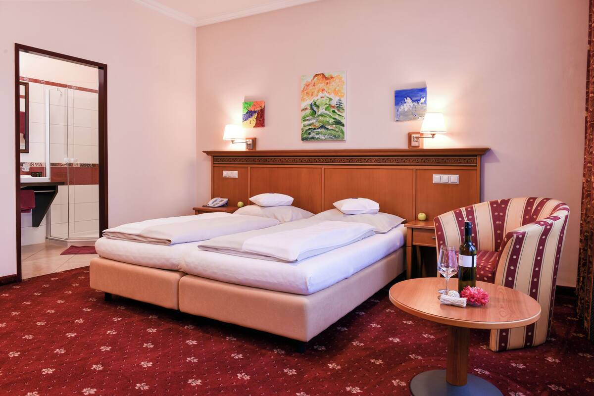 A hotel room with a double bed, a small round table with a bottle and glasses, a chair, and artwork on the wall.