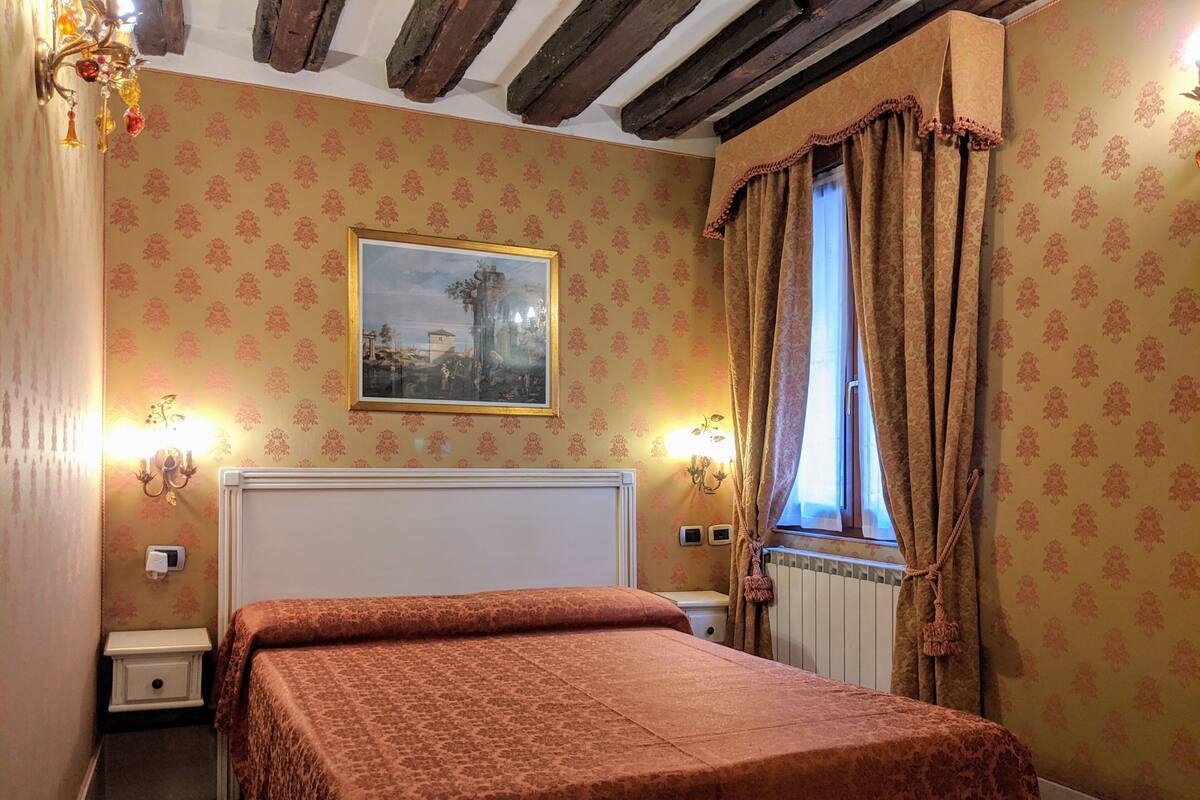 A bedroom with a bed, a nightstand, a wall-mounted lamp, a window with curtains, and a framed picture on the wall.