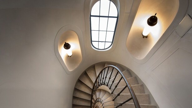 Un escalier en colimaçon doté d’une rampe centrale, flanqué de deux appliques murales éclairées et surmonté d’une lucarne.
