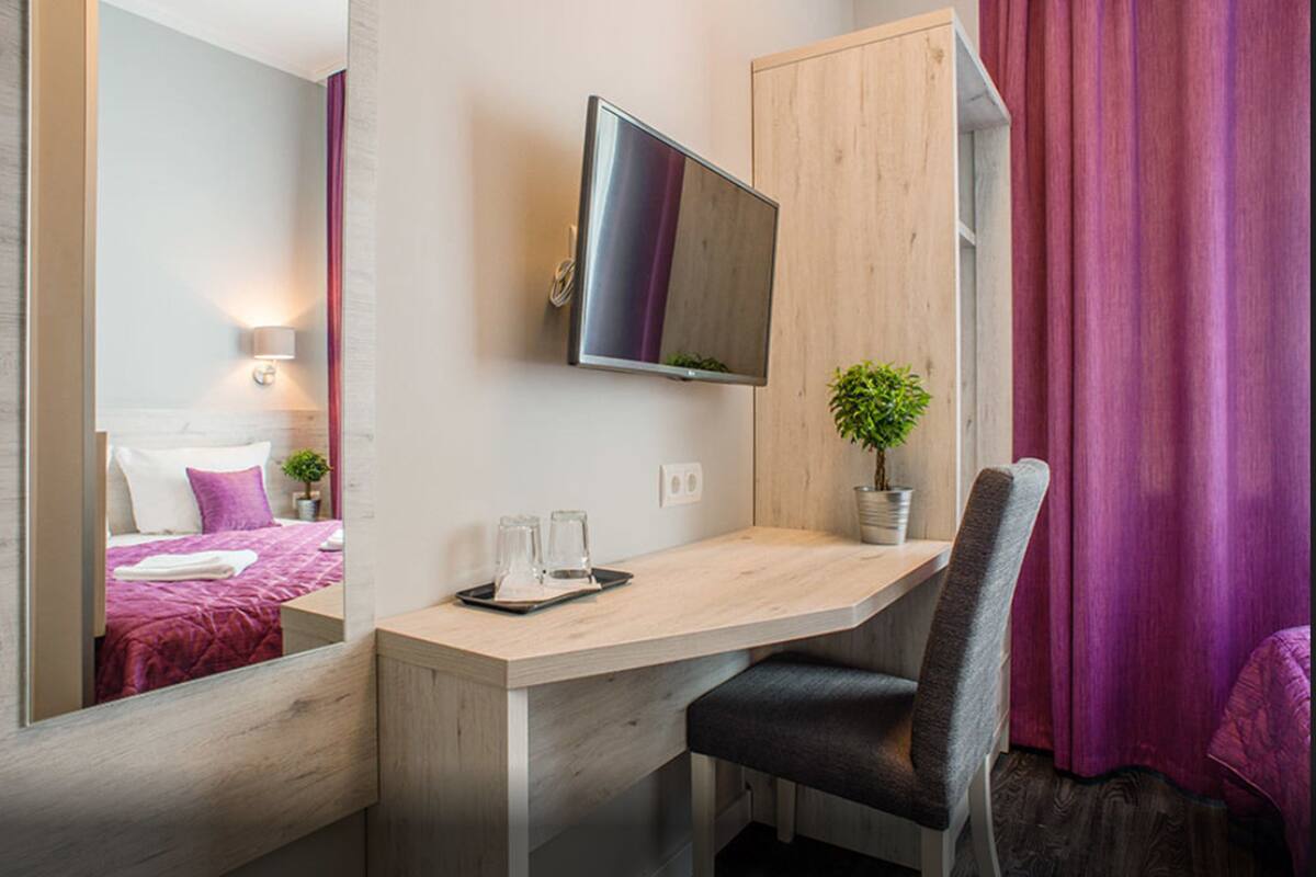 A hotel room with a wooden desk, a flat-screen TV, a potted plant, and a bed with purple bedding.