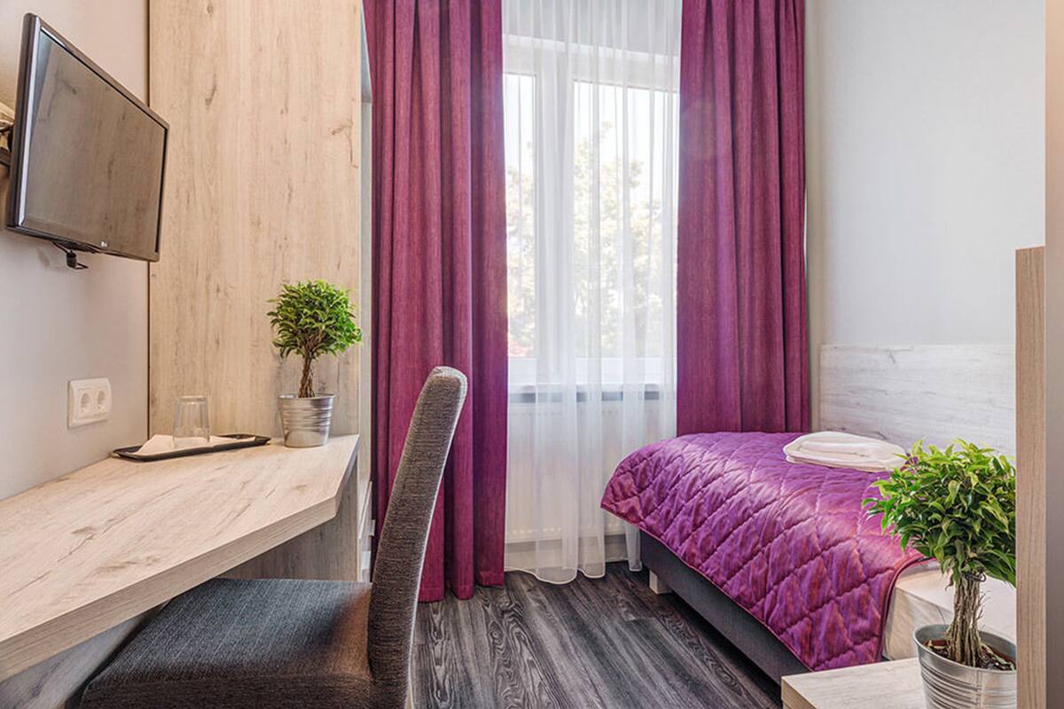 A bedroom with a bed, a desk, a chair, a TV, and a window with curtains.