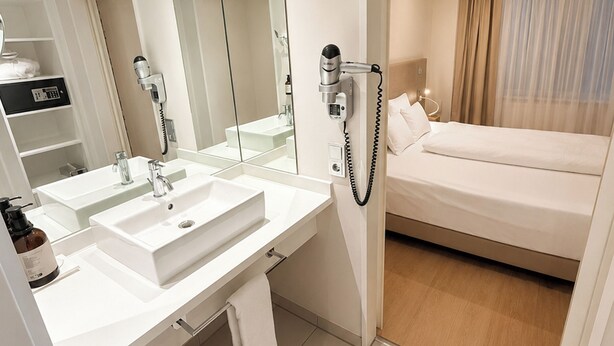 A hotel room with a large mirror, two sinks, a hairdryer, a bed, and a nightstand.