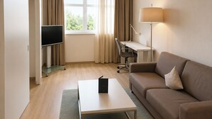 A modern hotel room with a flat-screen TV, a desk with a chair, a sofa, a lamp, and a window with curtains.