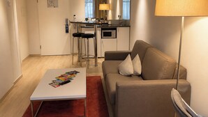 A modern living room with a sofa, a coffee table, and a lamp. There is a kitchen area with a microwave and a bar stool.