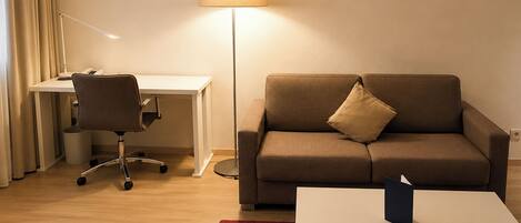 A hotel room with a desk, chair, sofa, lamp, and a small table.