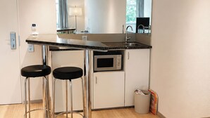 A compact kitchen with a countertop, two stools, a microwave, and a trash bin.