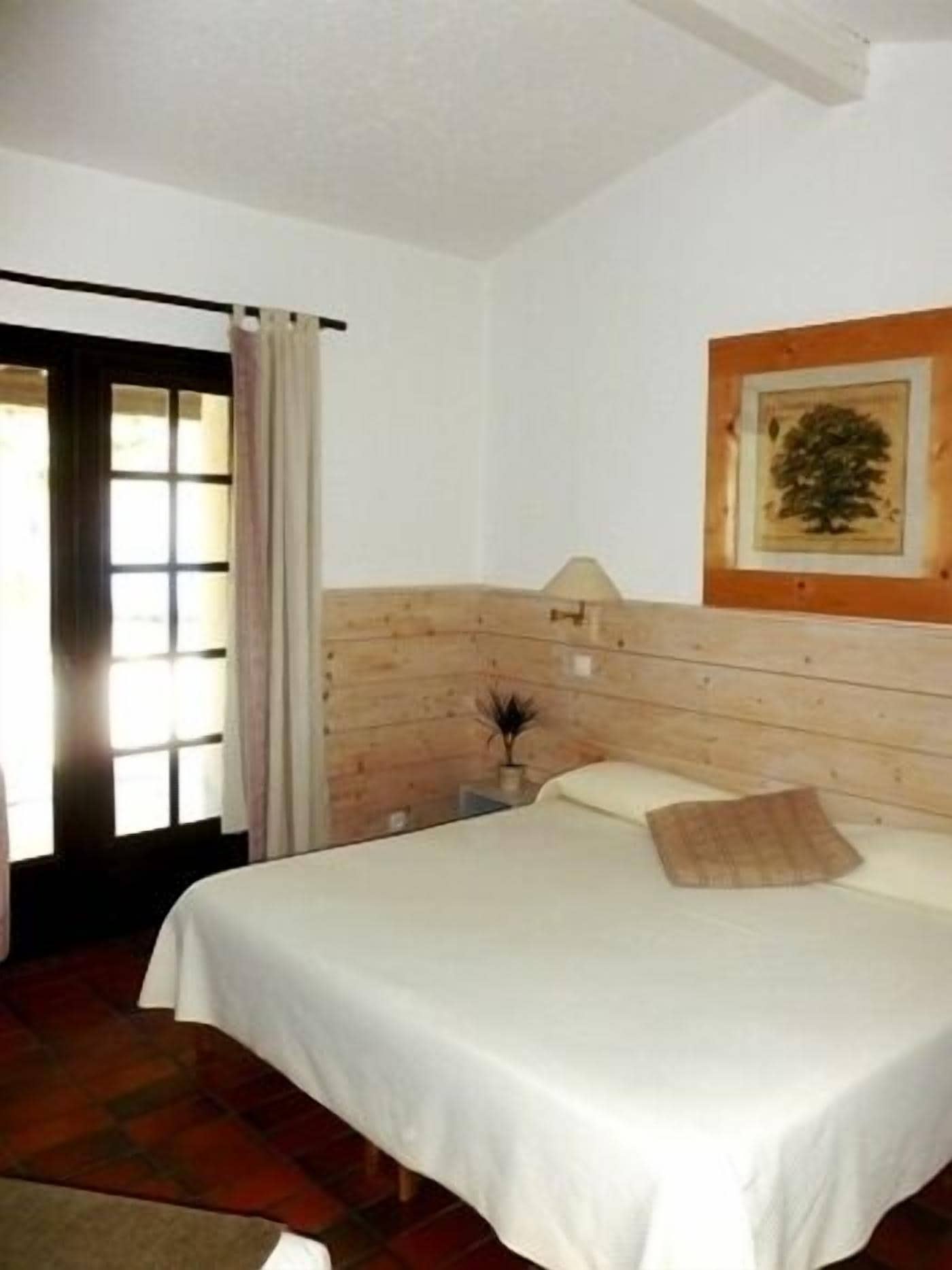 Une chambre à coucher avec une tête de lit en bois, un lit recouvert de draps blancs, un tableau encadré au mur et une fenêtre avec des rideaux.