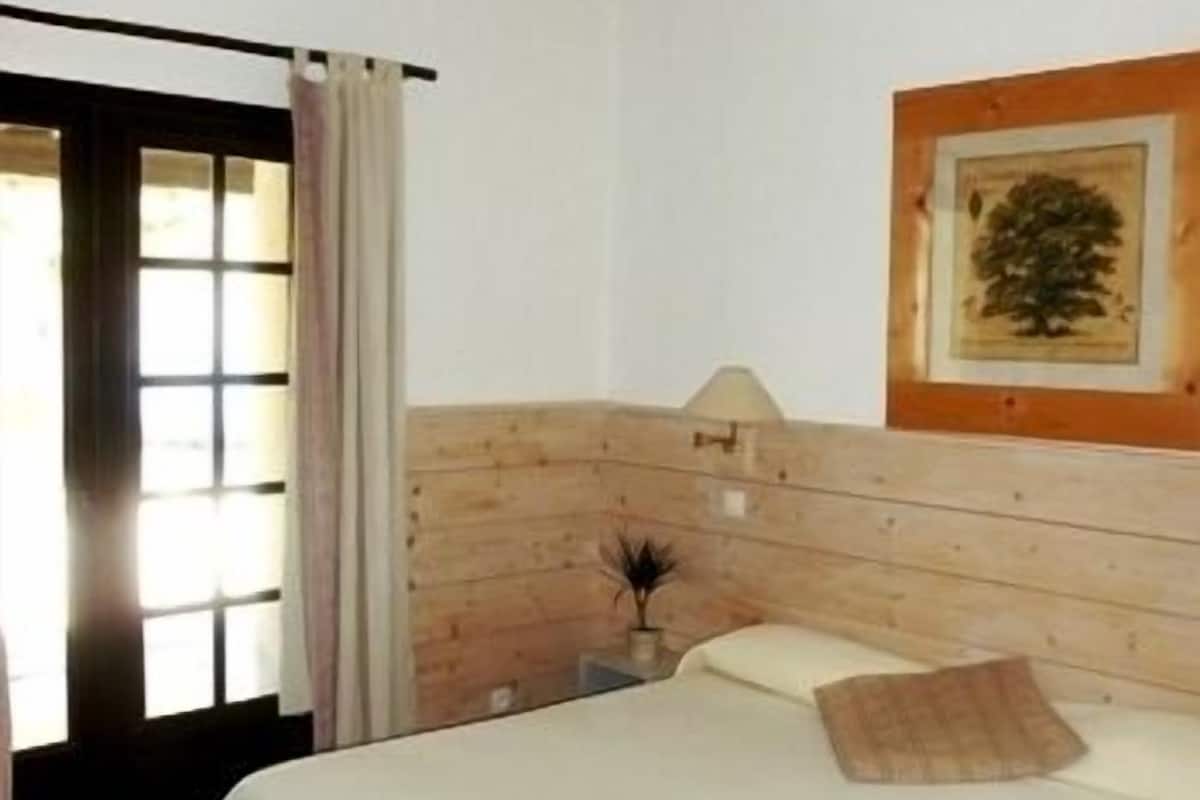 Ein Schlafzimmer mit einem hölzernen Kopfteil, einem Bett mit weißer Bettwäsche, einem gerahmten Bild an der Wand und einem Fenster mit Vorhängen.