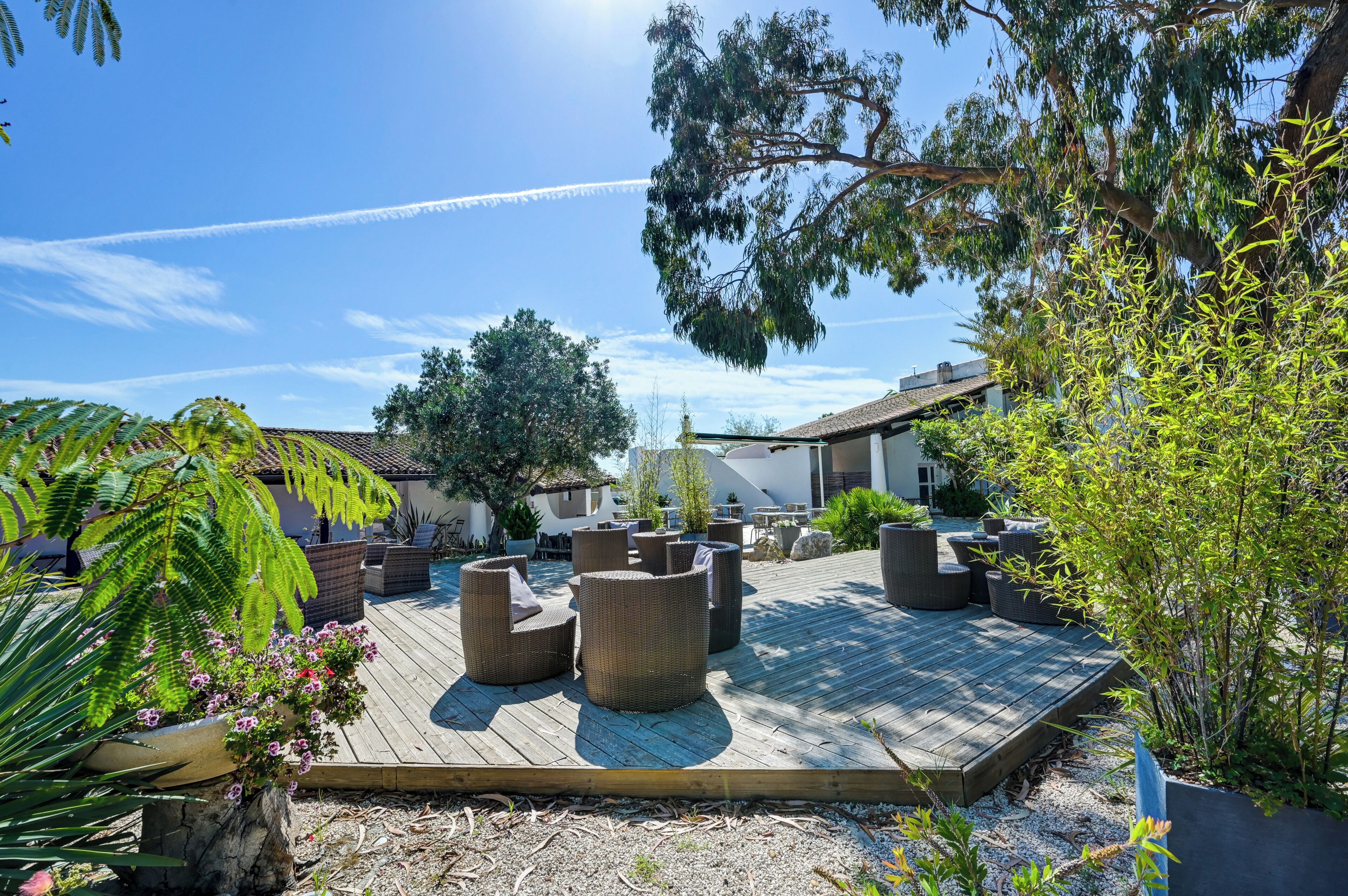 Une terrasse en bois, meublée de meubles en osier, entourée de verdure et sous un ciel dégagé.
