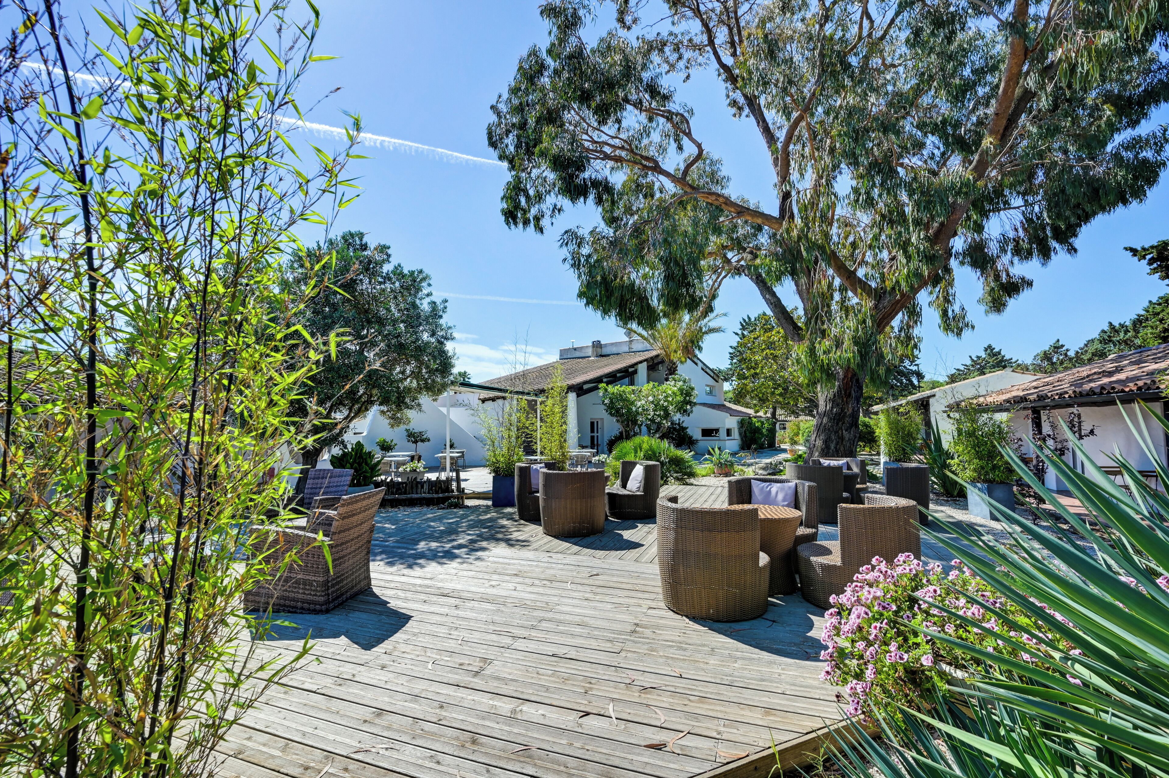 A wooden deck with wicker furniture, surrounded by greenery and a building with a tiled roof.