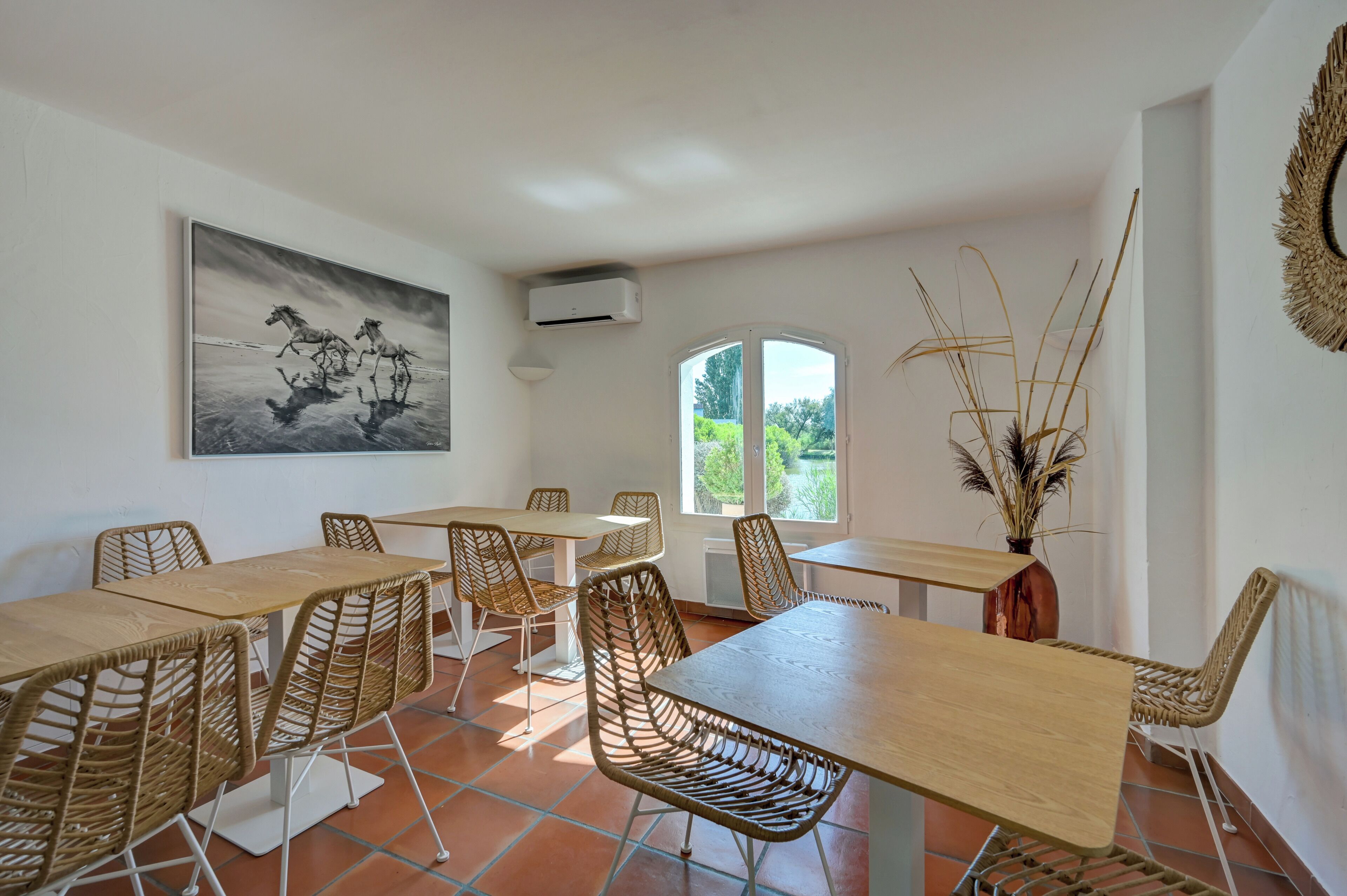 A dining area with wicker chairs, a wooden table, a painting of horses, a window, and a vase with plants.