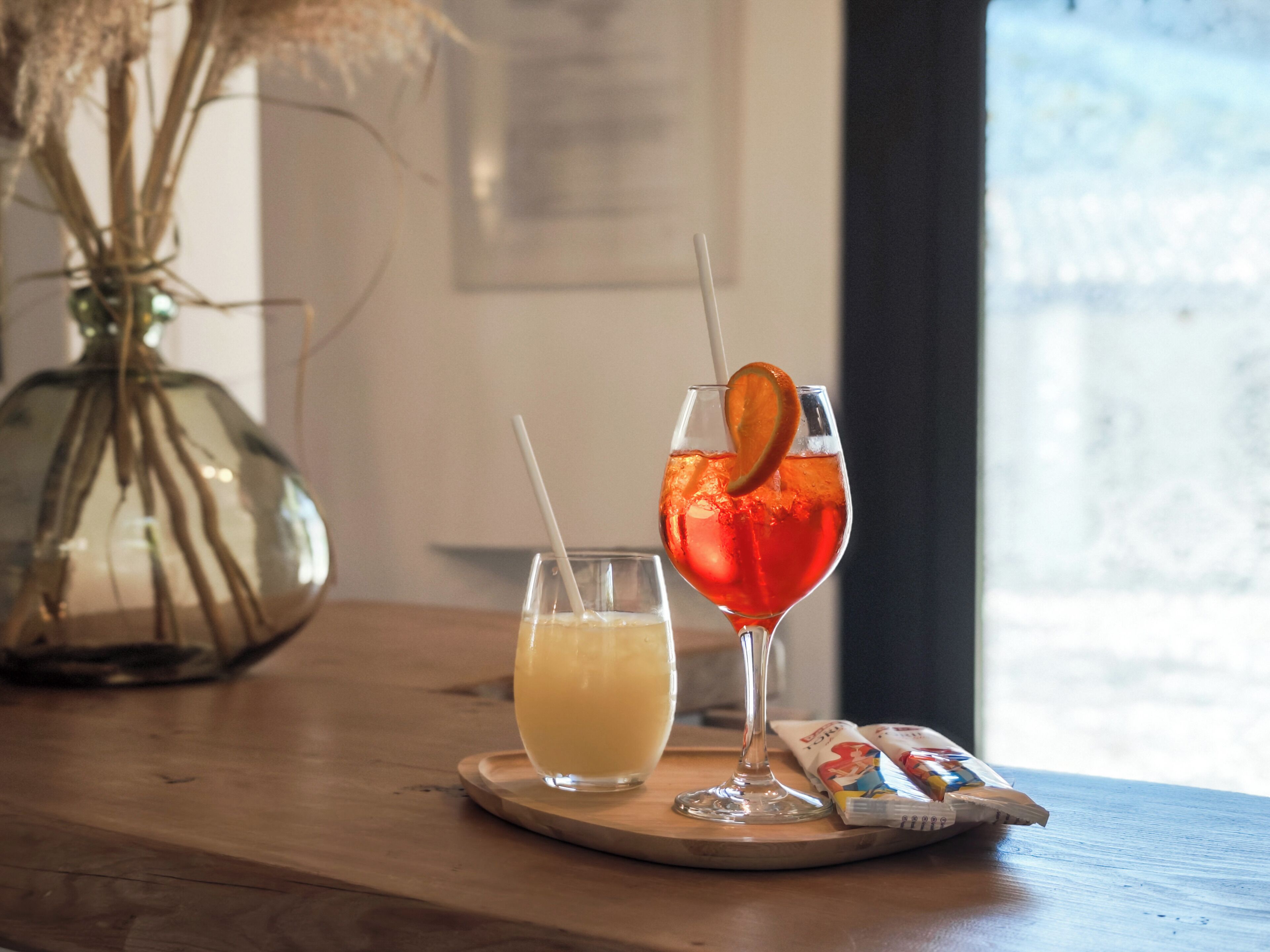 Two drinks on a wooden table, a vase with dried plants, and a window with a view.