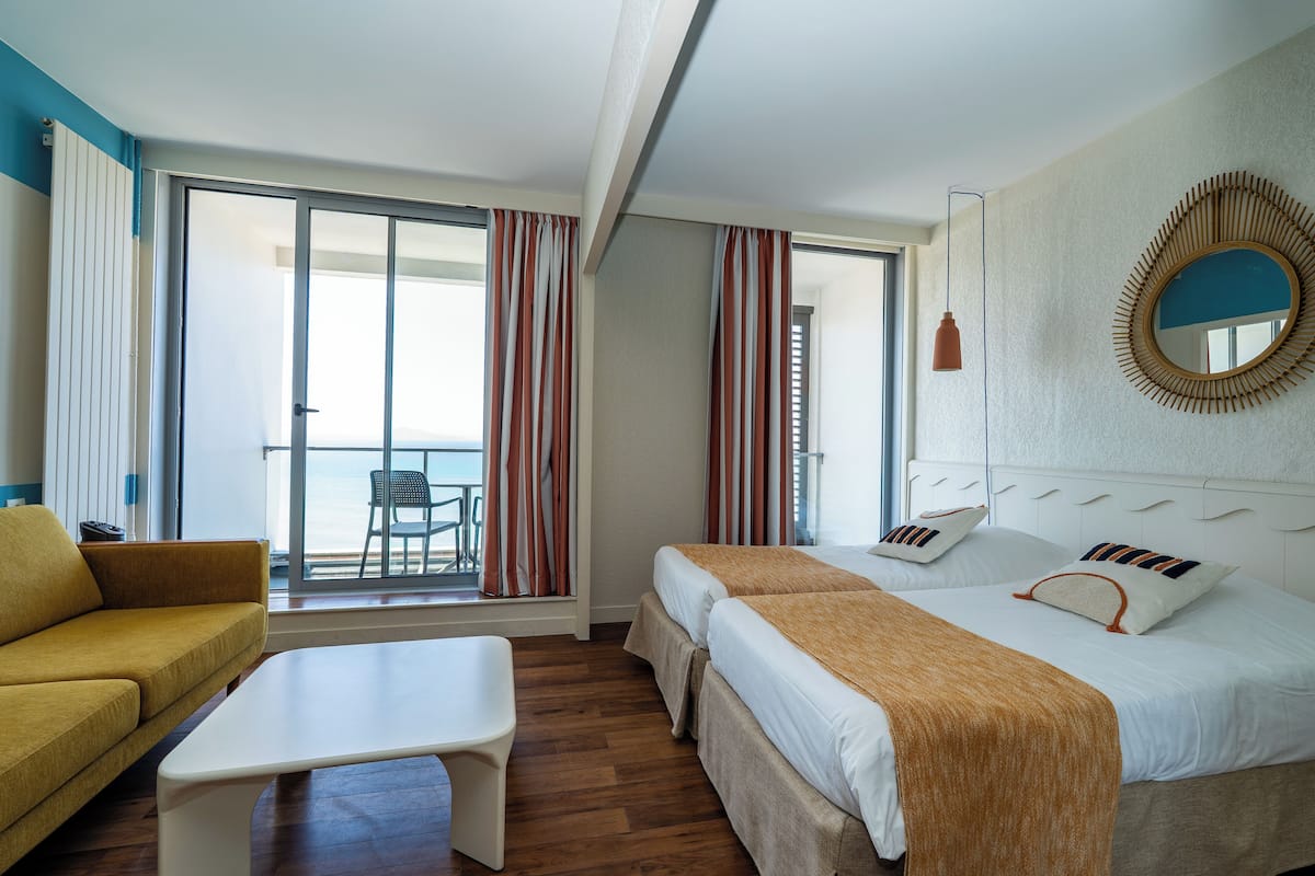 A hotel room with two beds, a sofa, a small table, and a balcony with curtains.