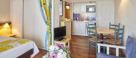 Habitación de hotel con cama, televisor, zona de comedor con mesa y sillas, cocina con armarios y microondas, y baño con inodoro y lavabo.