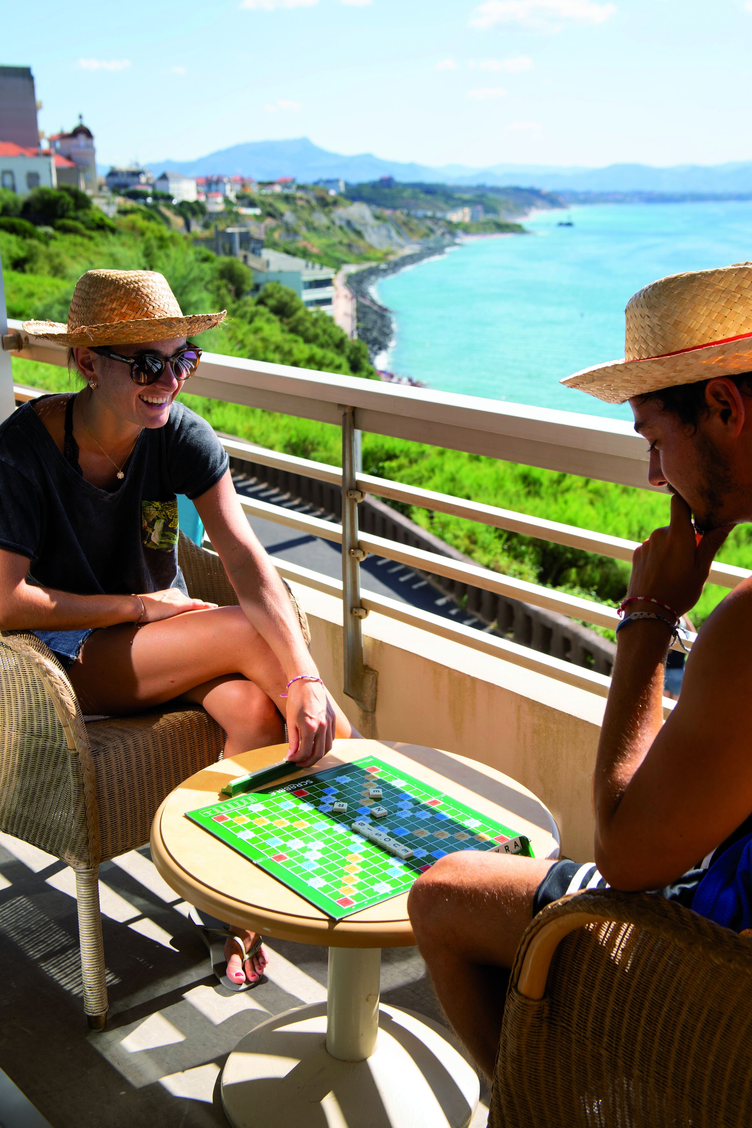 Two people playing a board game on a balcony overlooking a coastal area with houses and a view of the sea.