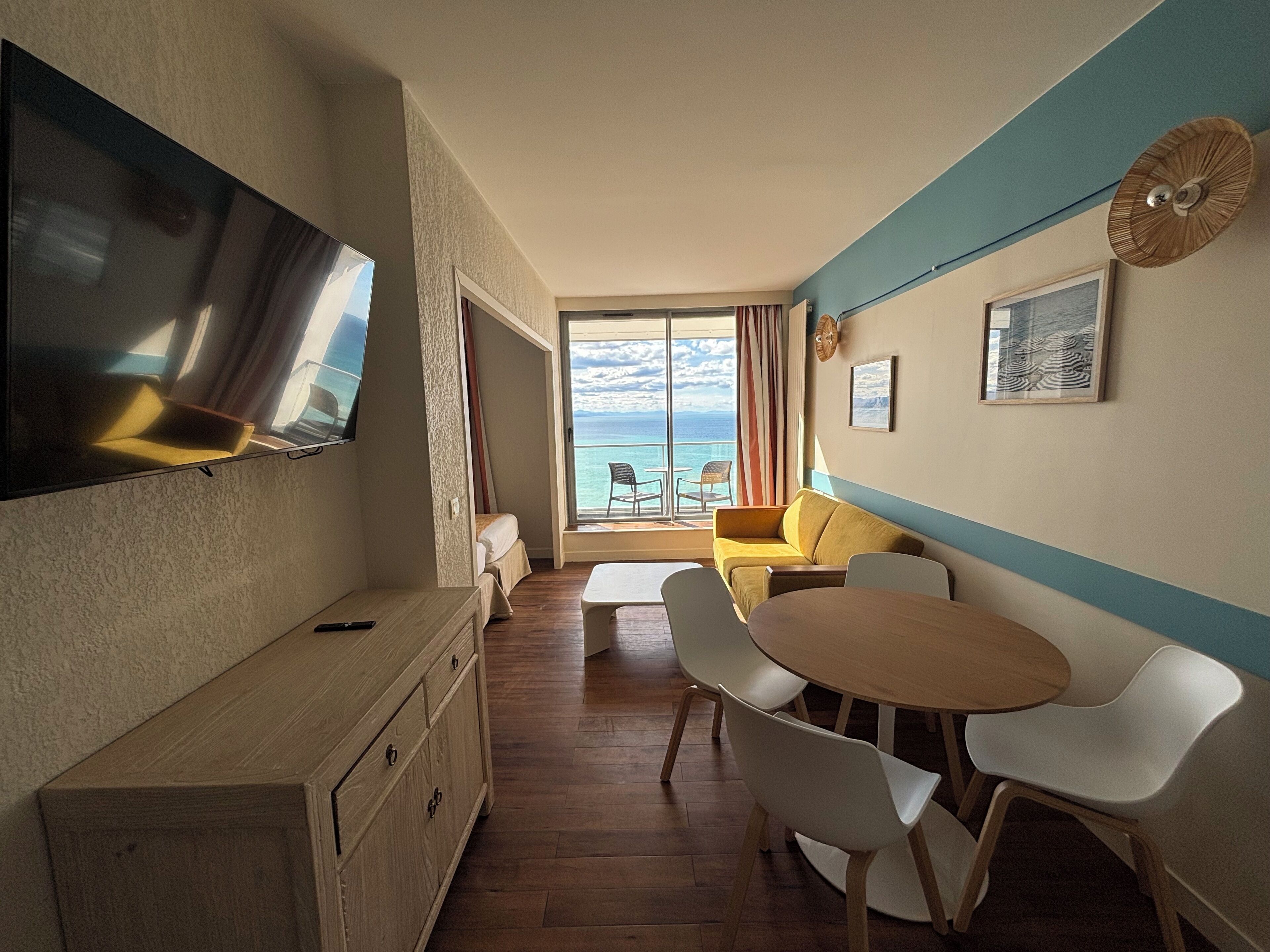 Een kamer met een aan de muur bevestigde televisie, een houten kast, een eettafel met stoelen en een bank. Er is een groot raam met uitzicht op de oceaan.