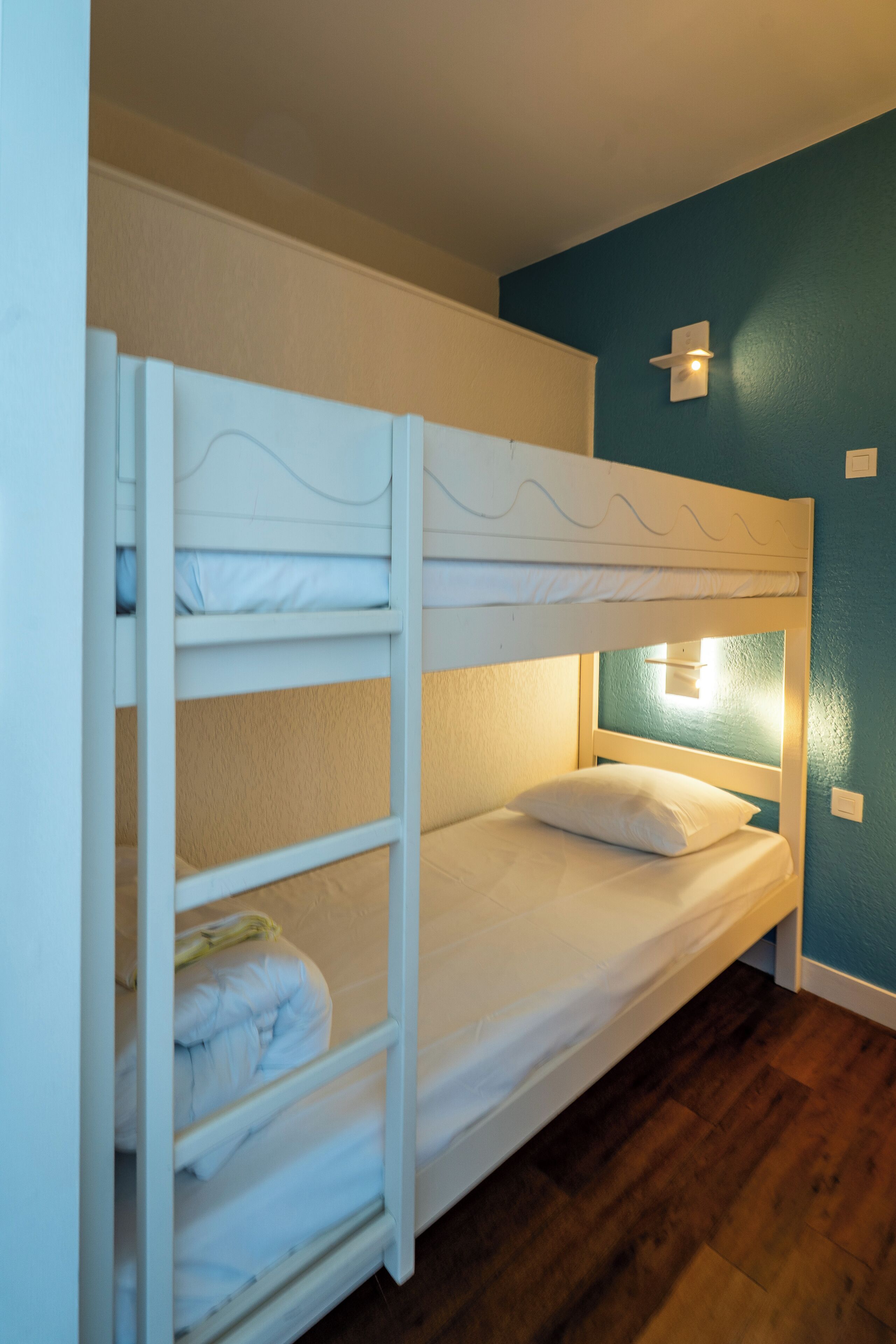 A bunk bed with a headboard light, wooden flooring, and a teal wall.