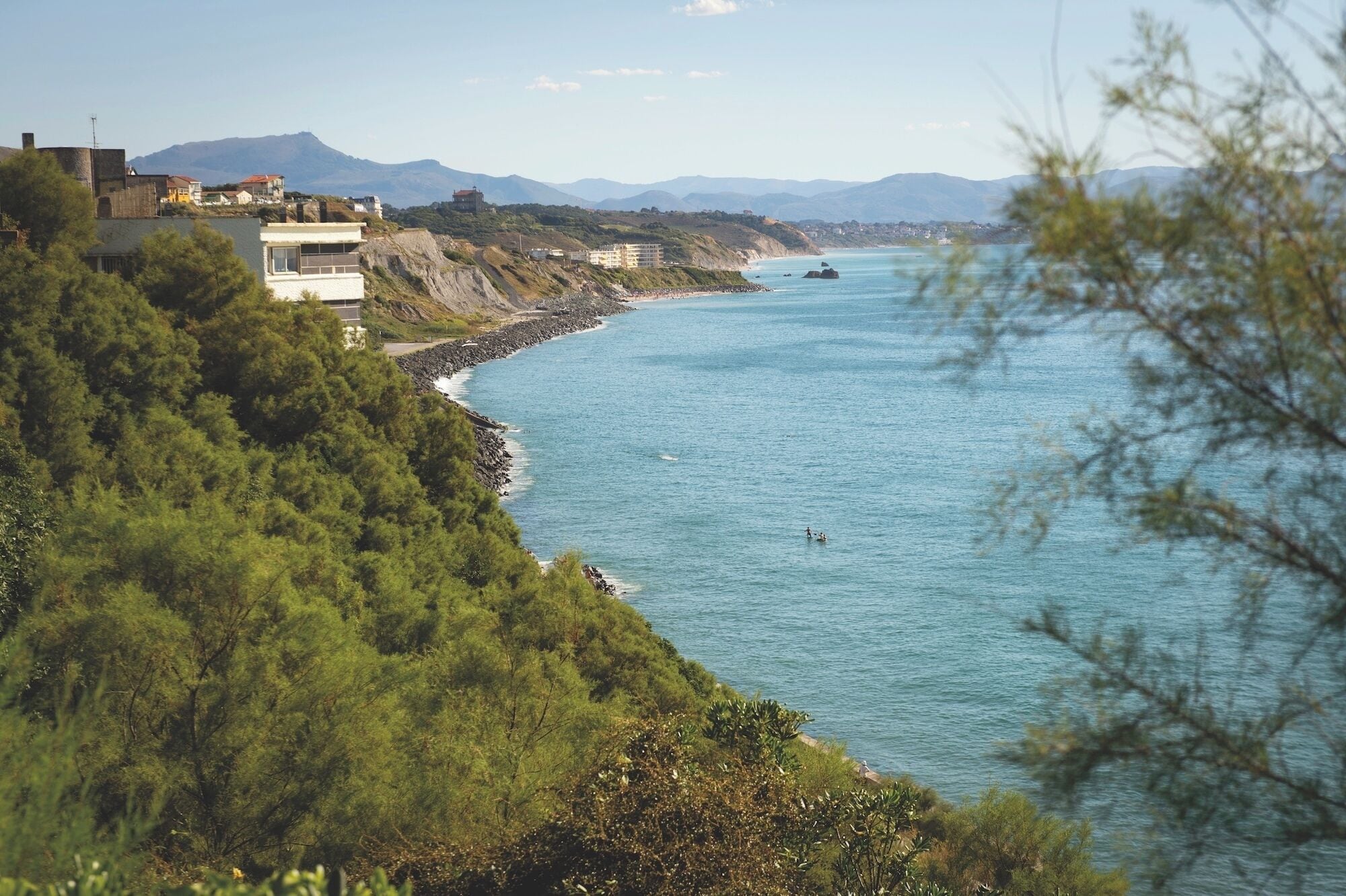 Vistes costaneres amb edificis, una riba rocosa i mar blau clar.