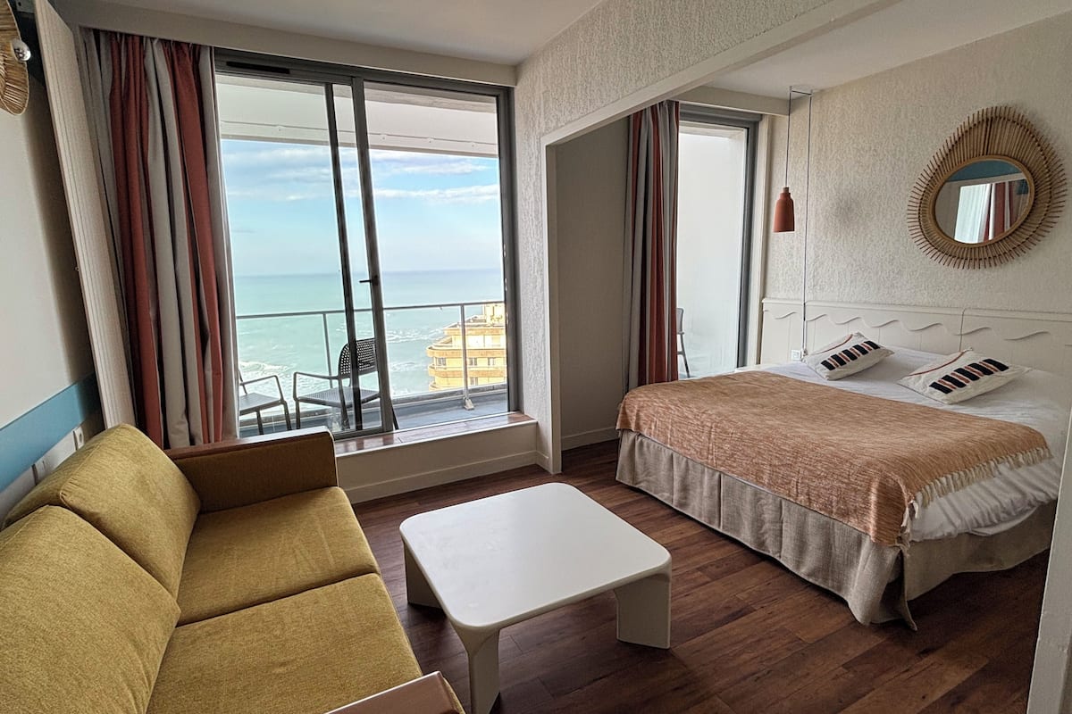 A hotel room with a bed, a sofa, a small table, and a view of the sea through large windows.