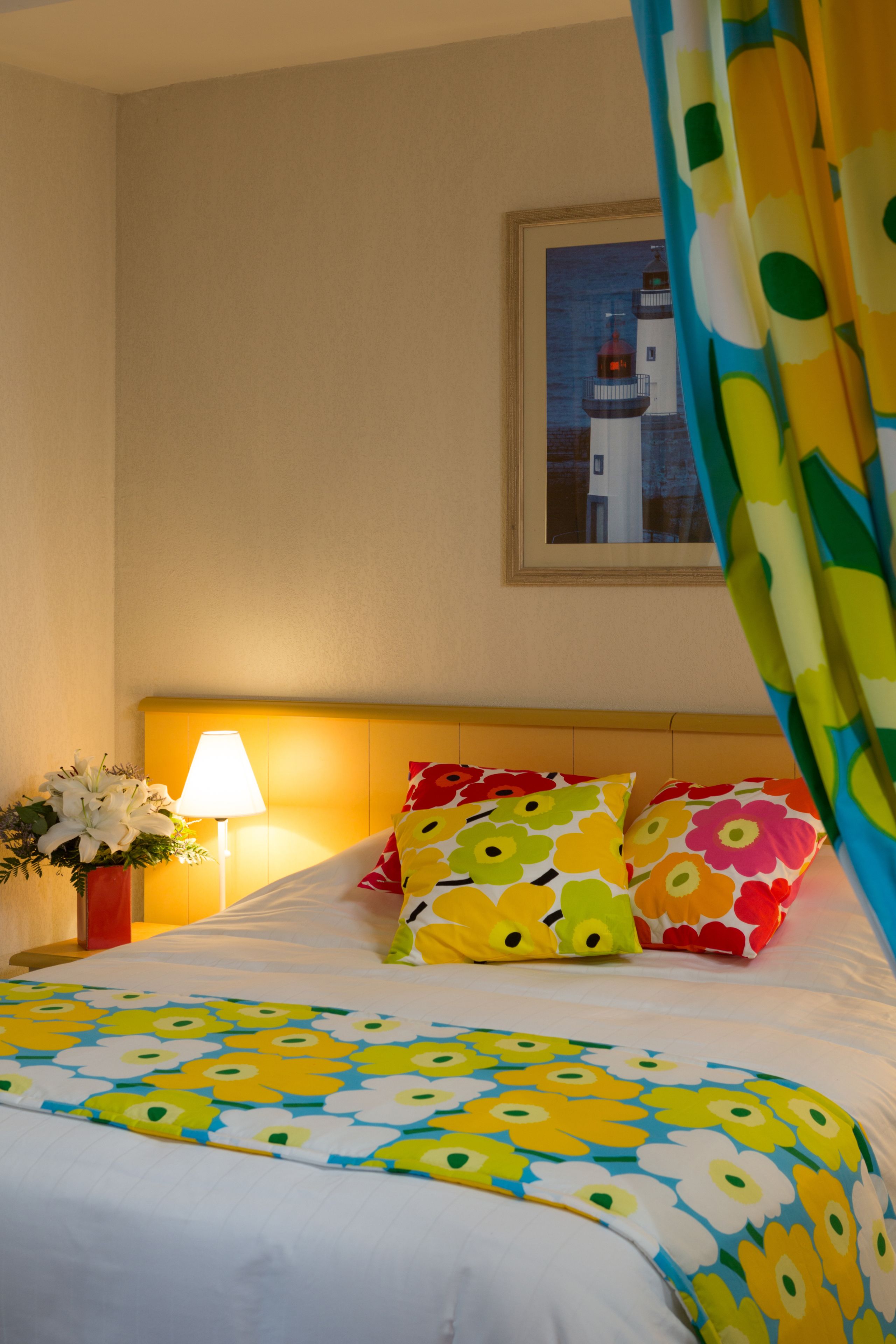 A hotel room with a bed, colorful pillows, a nightstand with a lamp, a vase with flowers, and a framed picture on the wall.