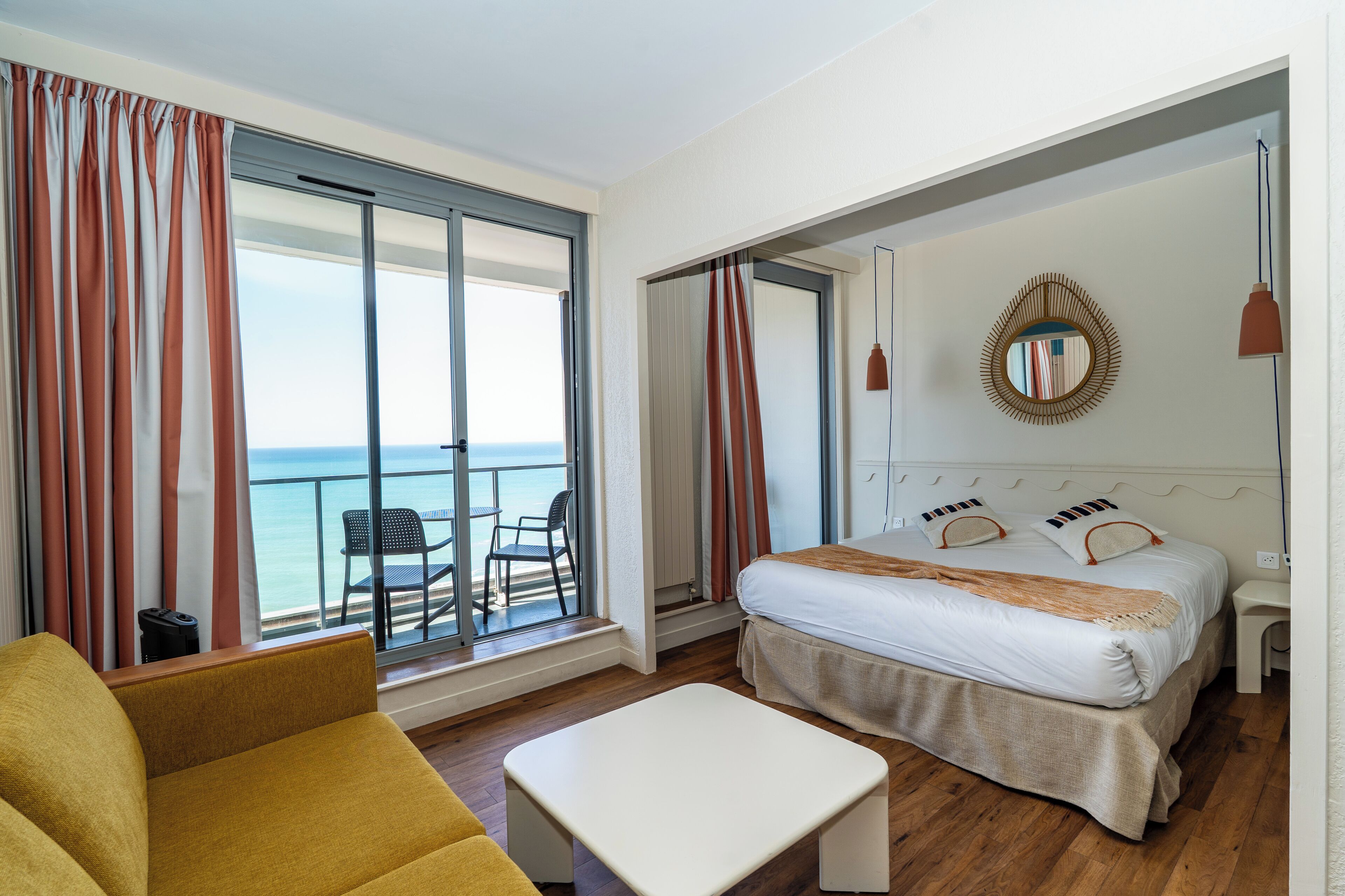 A hotel room with a bed, a sofa, a table, a balcony with a view of the sea, and a large window.