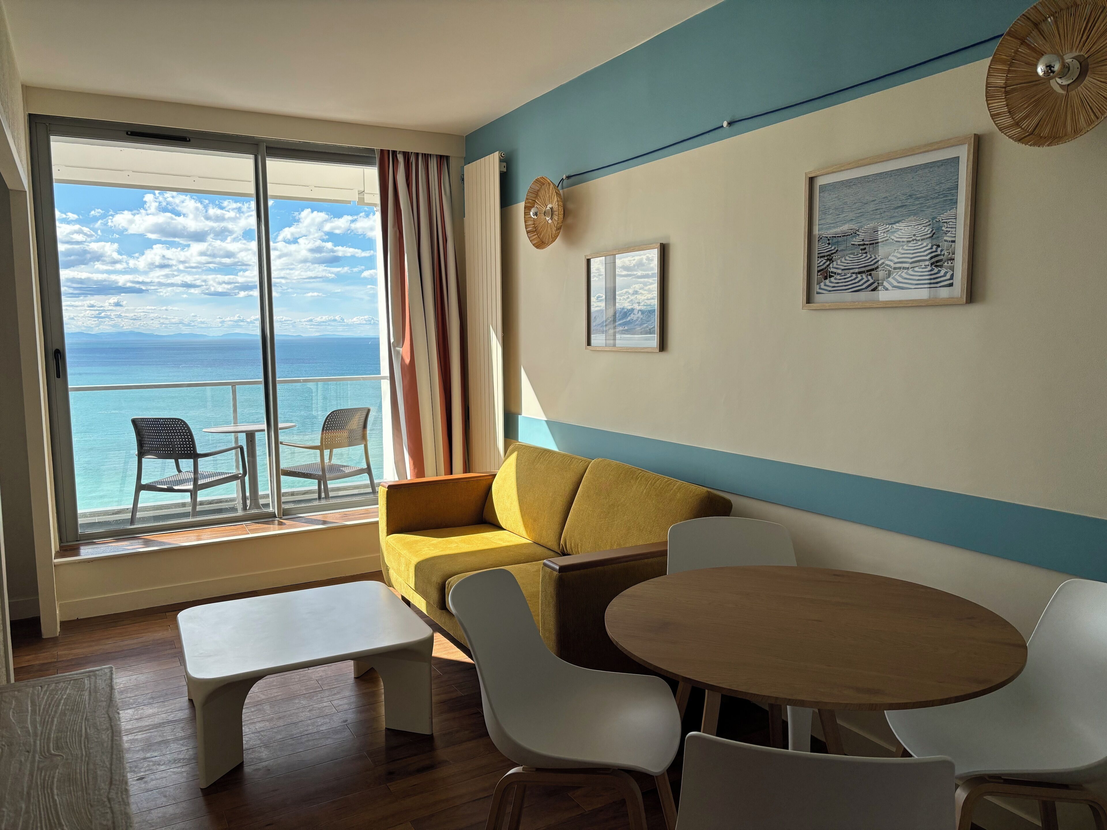 A room with a sofa, a round table, and two chairs. There is a window with a view of the ocean.