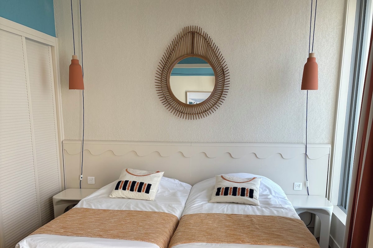 A double bed with beige bedding, a round mirror on the wall, and two pendant lights.