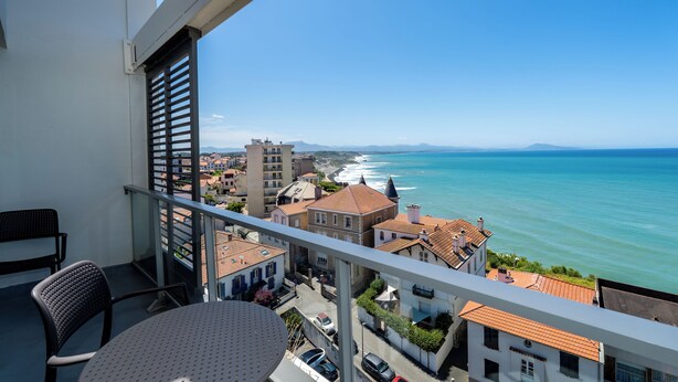 Un balcon avec une table et des chaises, donnant sur une ville côtière avec ses bâtiments et la mer.