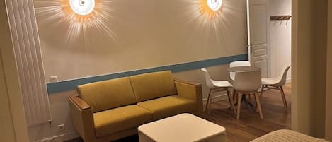 Habitación con un sofá, una mesita y dos sillas. Se observan dos luces fijadas a la pared y una puerta al fondo.