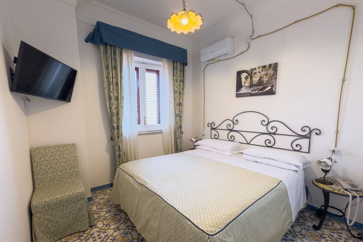 A bedroom with a bed, a chair, a TV, a window with curtains, an air conditioner, and a painting on the wall.