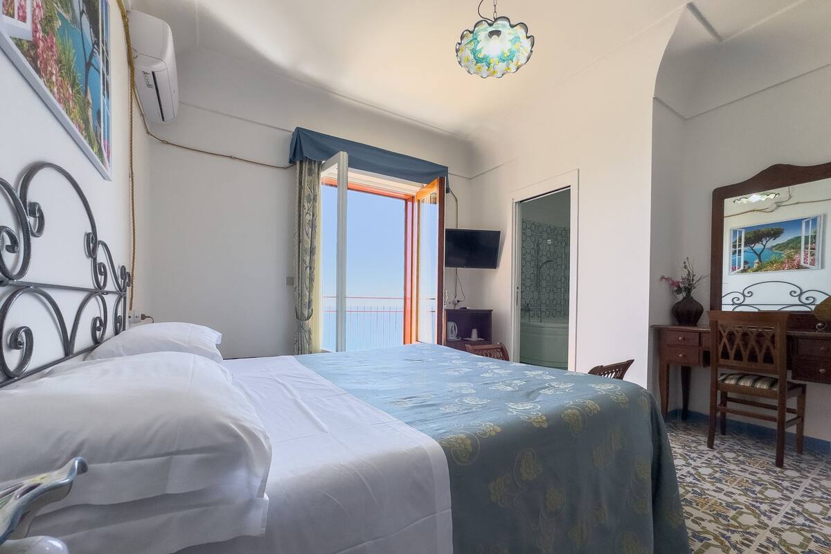 A hotel room with a bed, a desk, a chair, a TV, and a balcony with a view.