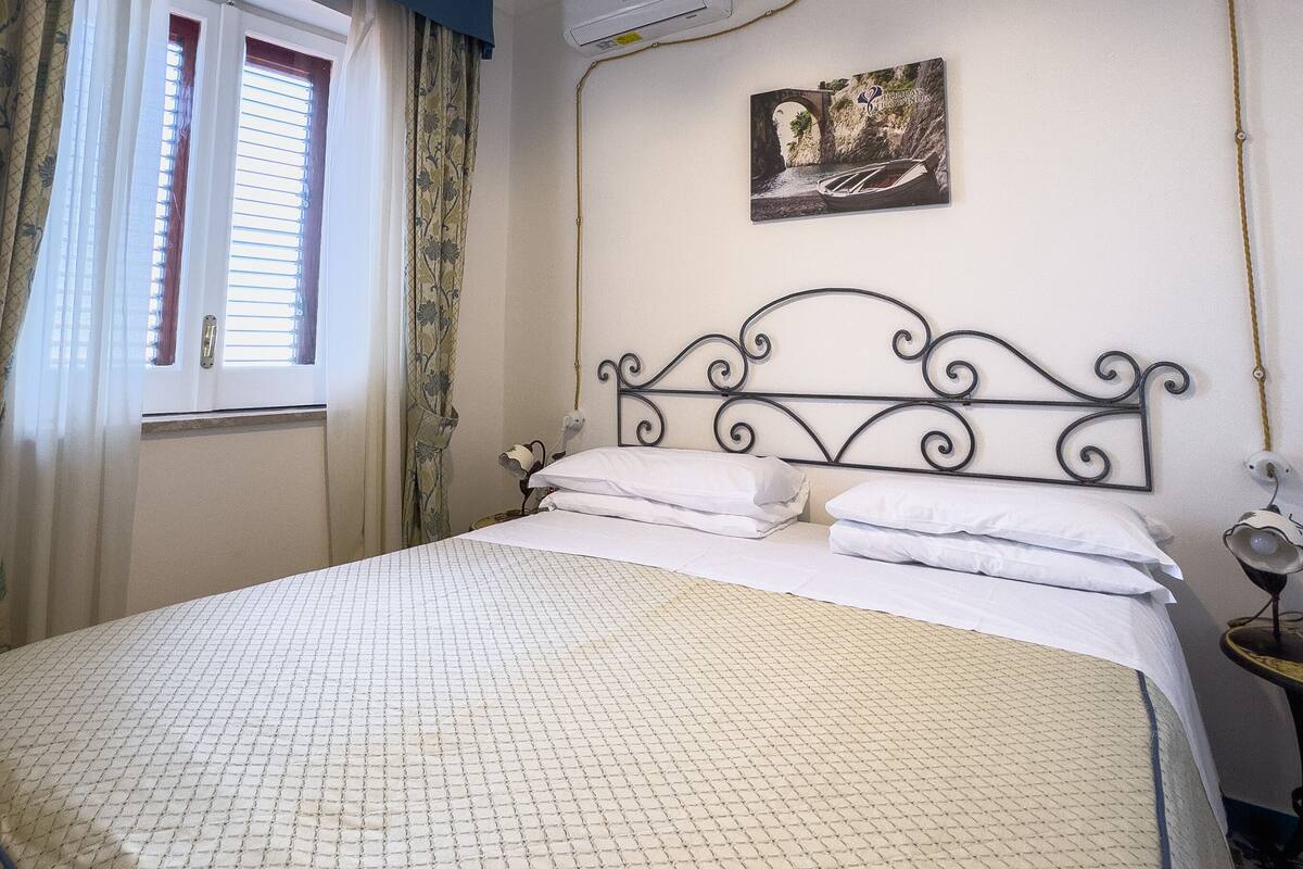 A bedroom with a bed, a window with curtains, an air conditioning unit, a wall-mounted picture, and a hanging light fixture.
