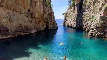 A clear coastal area with a stone bridge, rocky cliffs, and people swimming in the water.