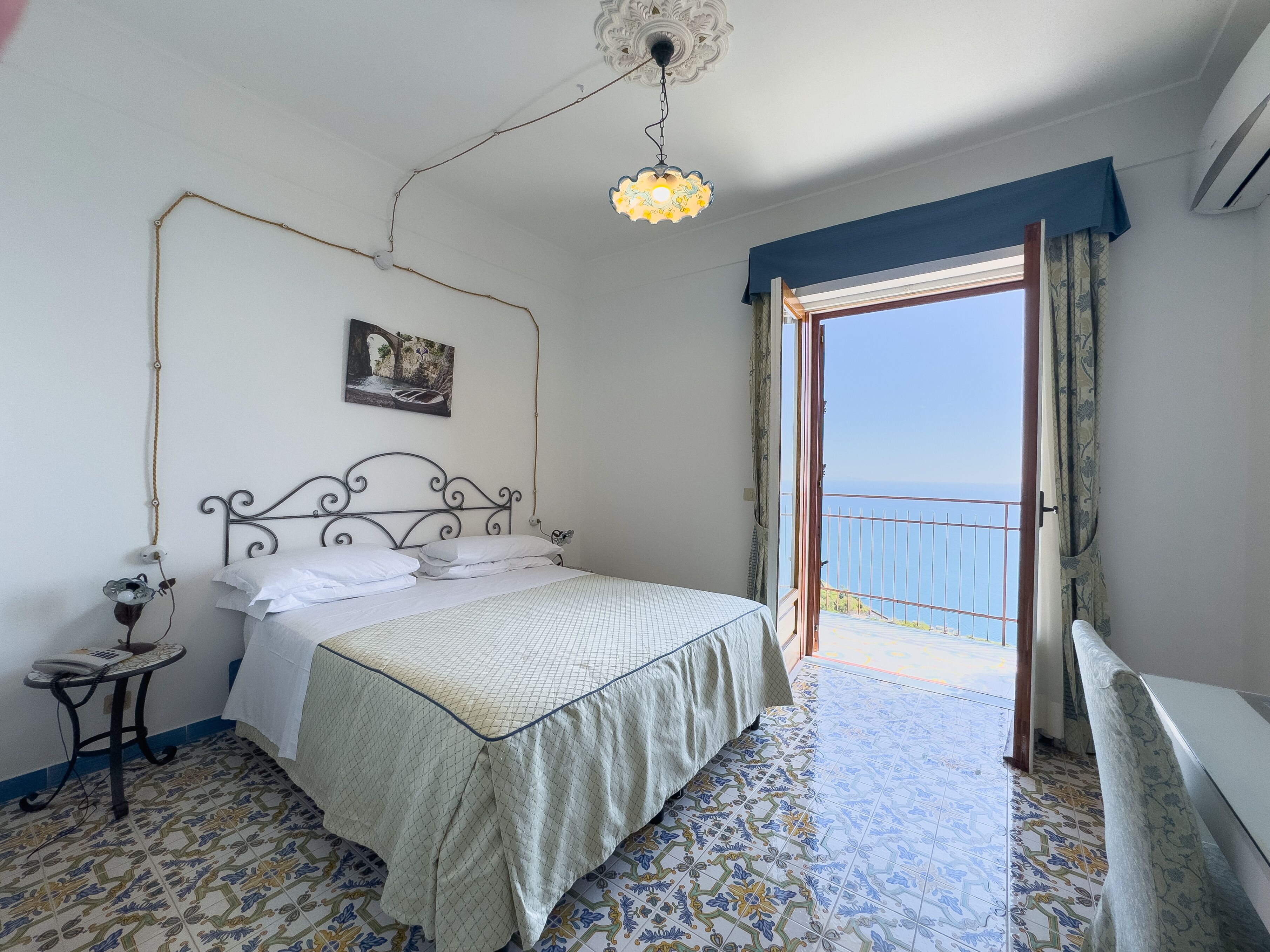 A bedroom with a bed, a bedside table, a hanging light fixture, a painting, and a view of the sea through an open door.