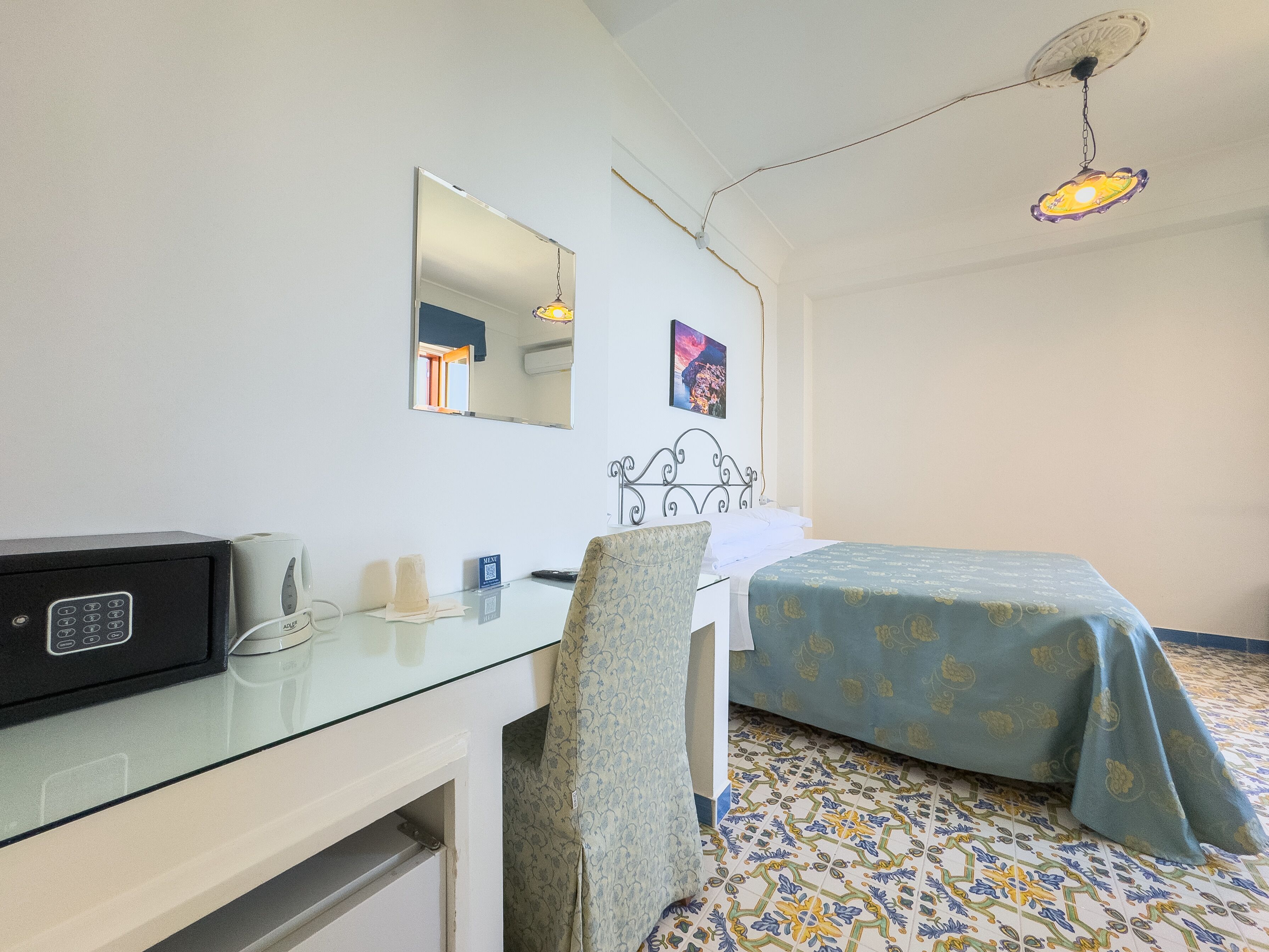 Una habitación de hotel con una cama, un escritorio con teléfono, una silla, un espejo y una lámpara fijada a la pared.