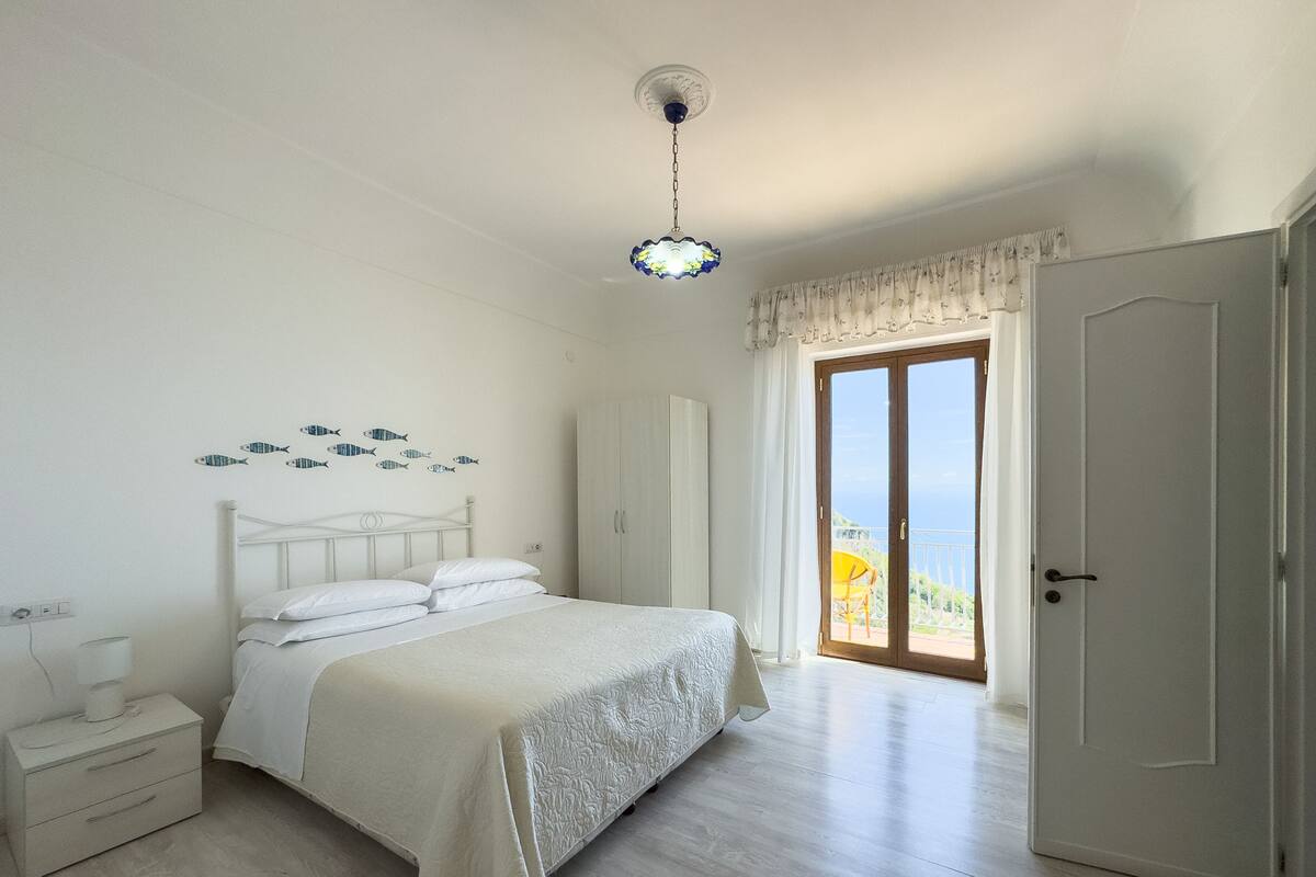 A bedroom with a bed, a nightstand, a dresser, a door leading to a balcony, and a ceiling light.