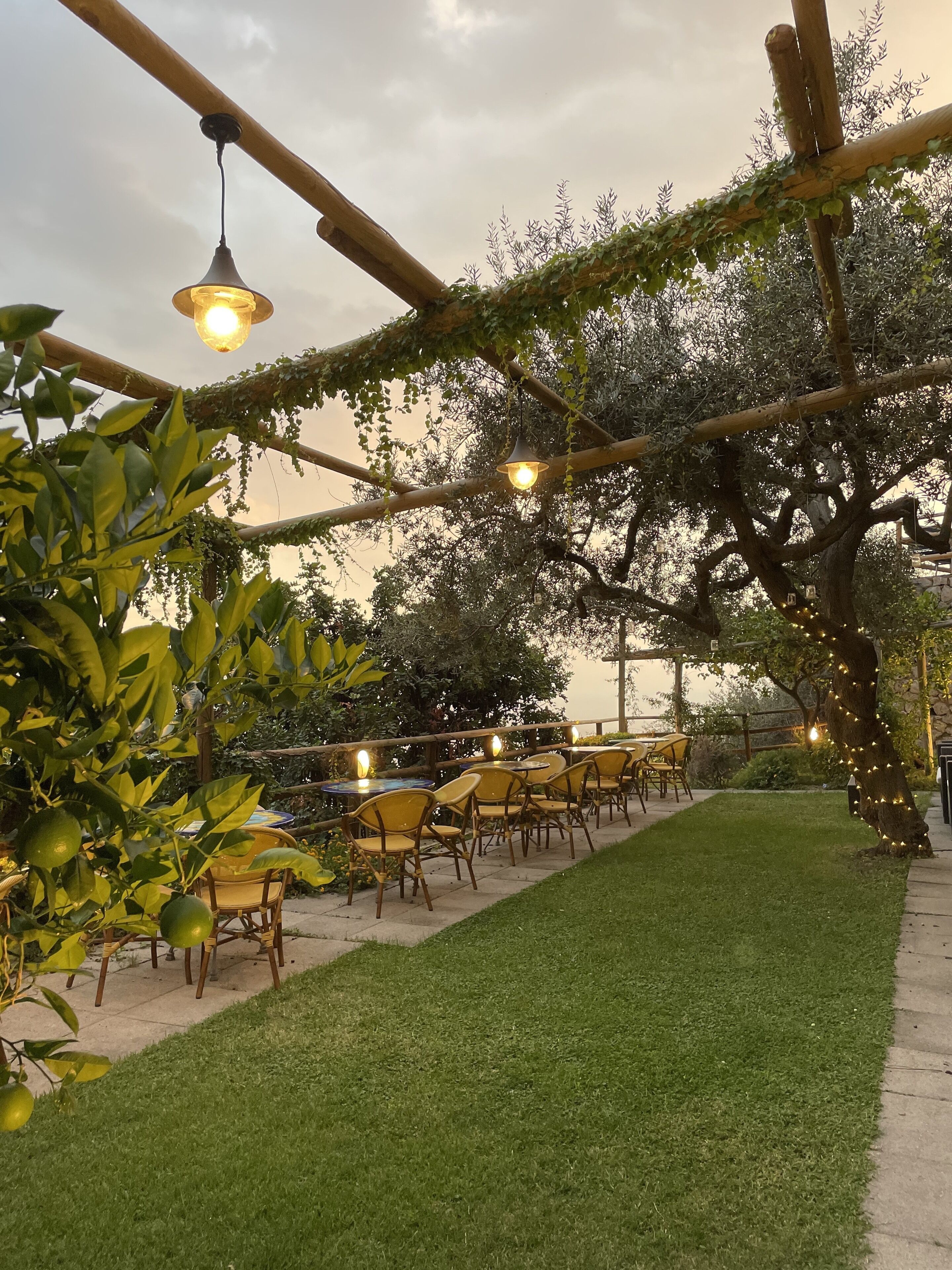 An outdoor seating area with tables and chairs under a pergola with hanging lights, surrounded by greenery and a well-maintained lawn.
