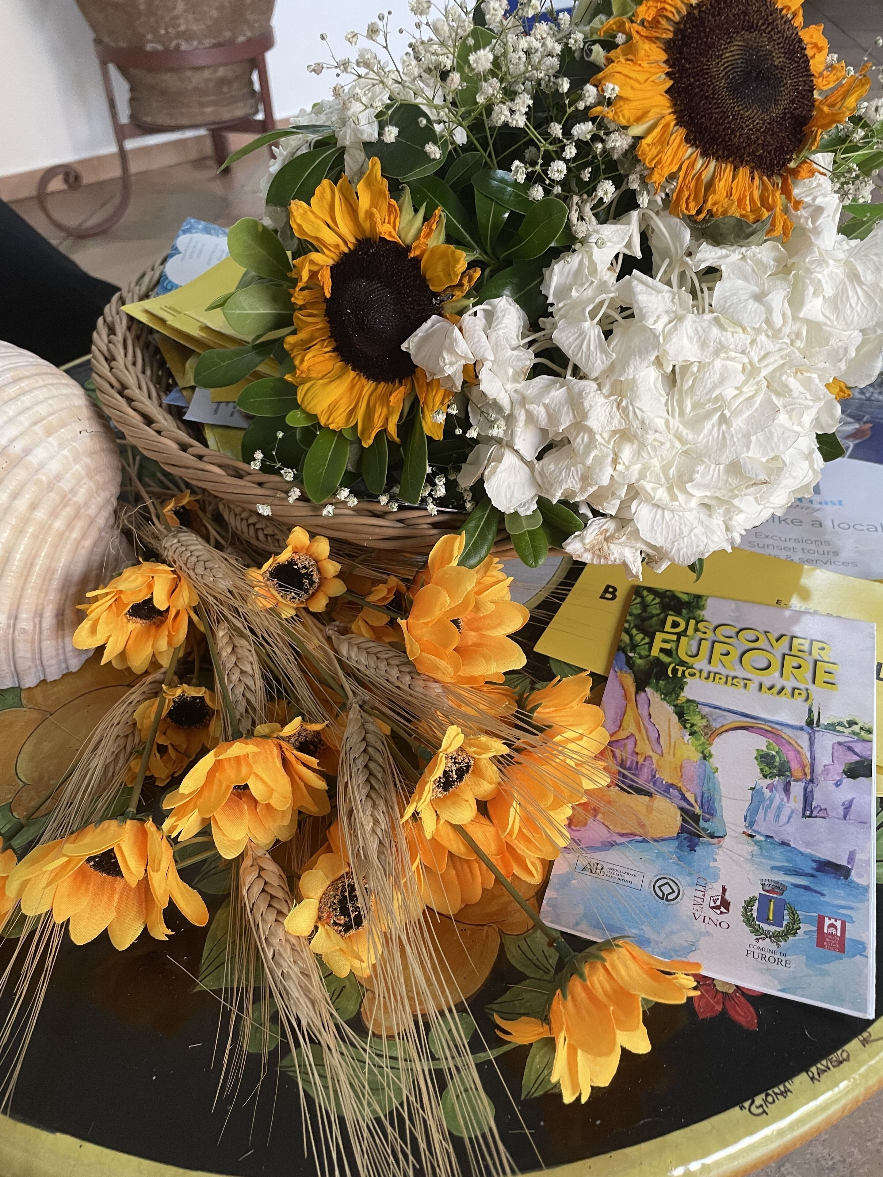 A table with a basket of sunflowers, white flowers, and wheat stalks, along with a tourist map of Furore.