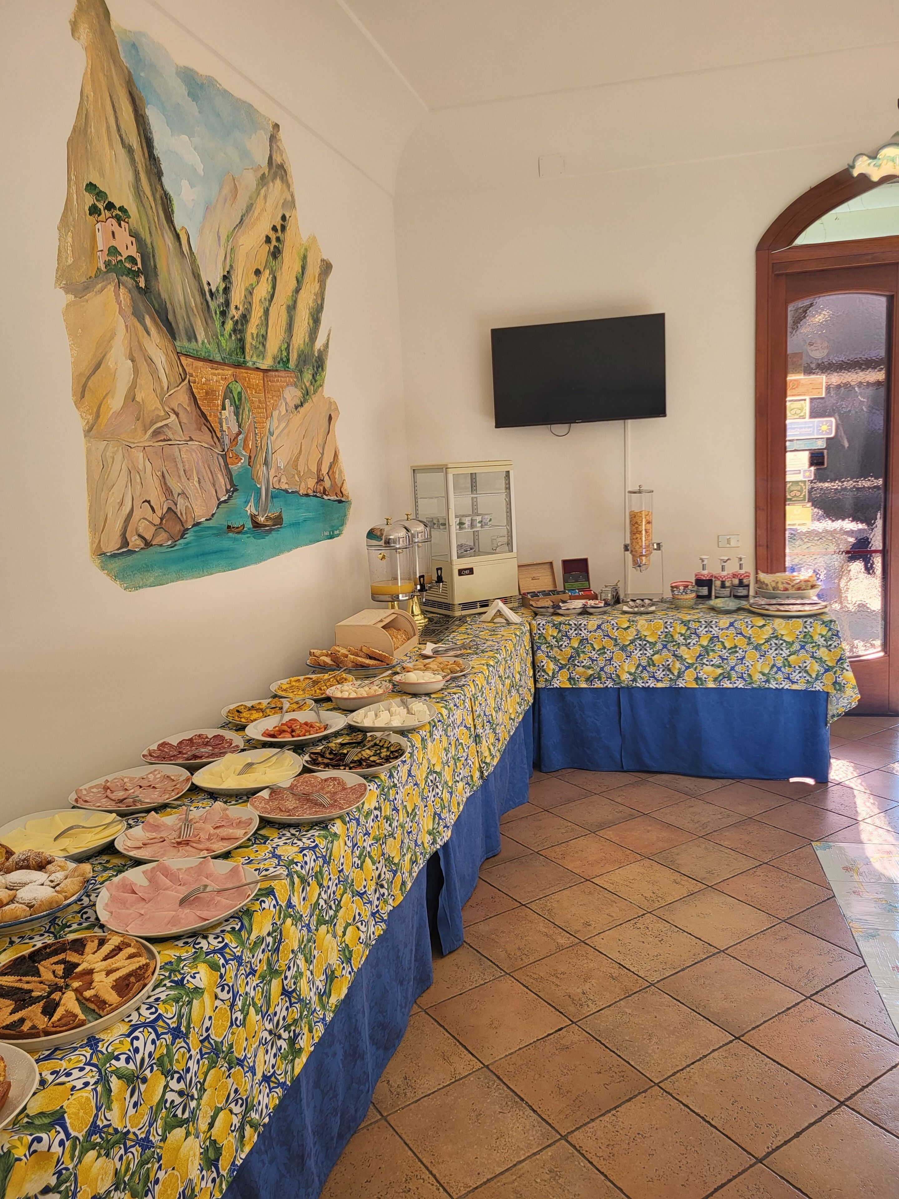 A buffet setup with various dishes, a painting of a coastal scene, and a television on the wall.