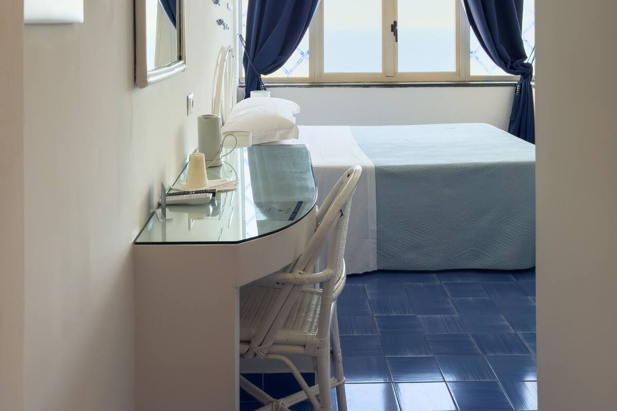 A bedroom with a bed, a bedside table, a mirror, a window with curtains, and a ceiling light.