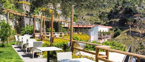 Een terras met tafels en stoelen, omgeven door begroeiing en met uitzicht op een heuvel.