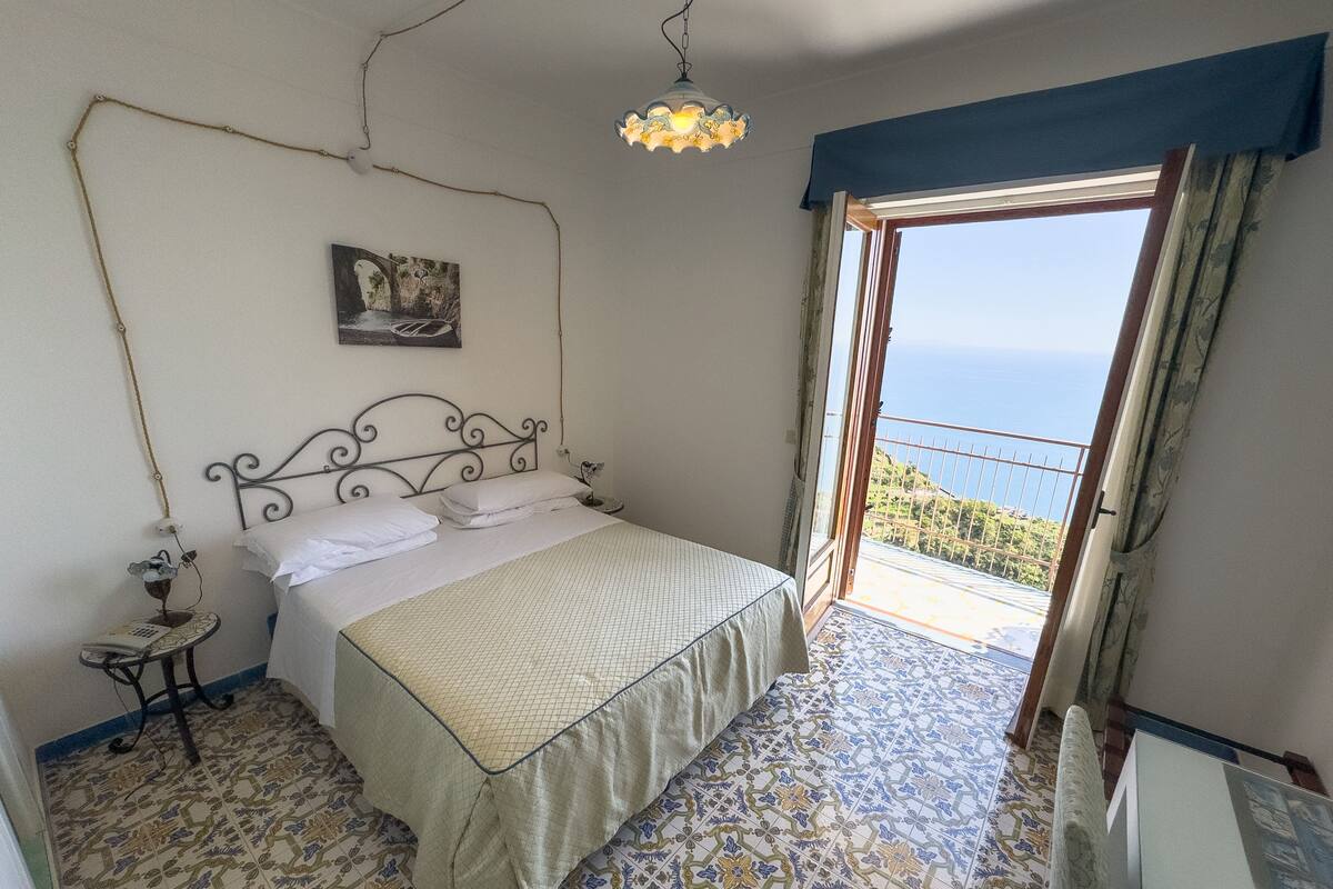 A bedroom with a bed, a nightstand, a wall-mounted lamp, a painting, and a view through an open door.