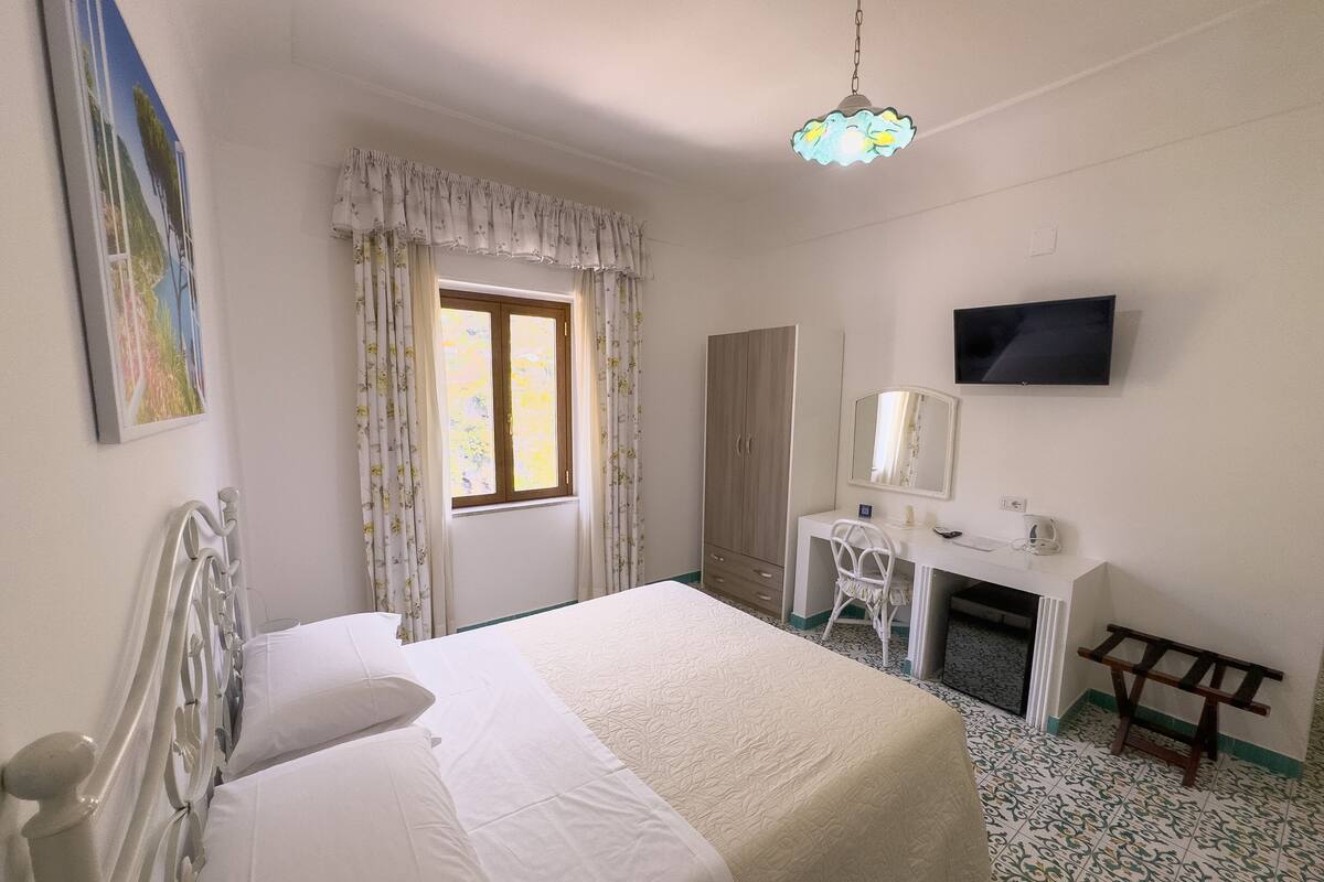 A bedroom with a bed, a desk, a chair, a wardrobe, a television, and a window with curtains.