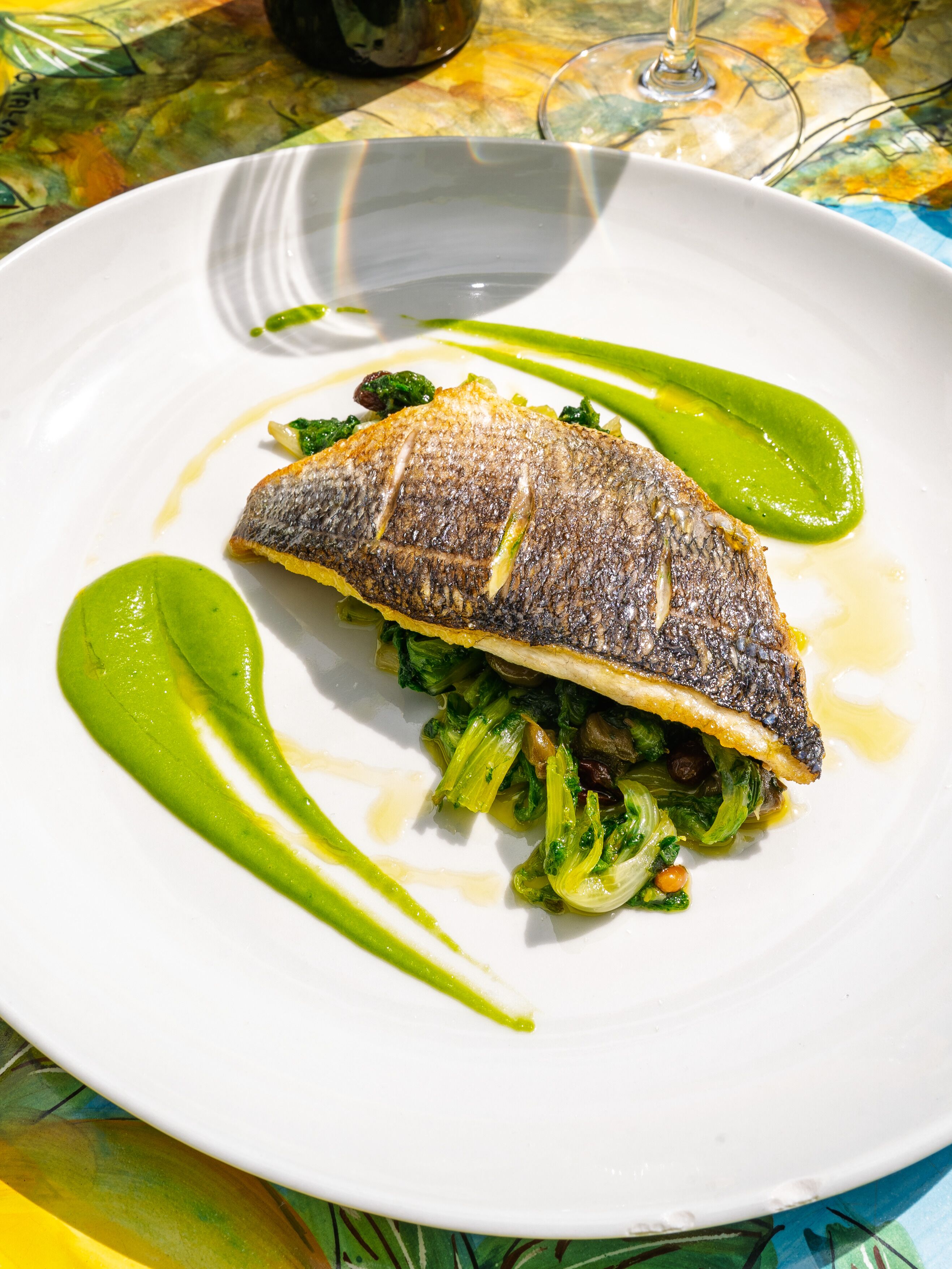 A white plate with a fish, greens, and a green sauce.